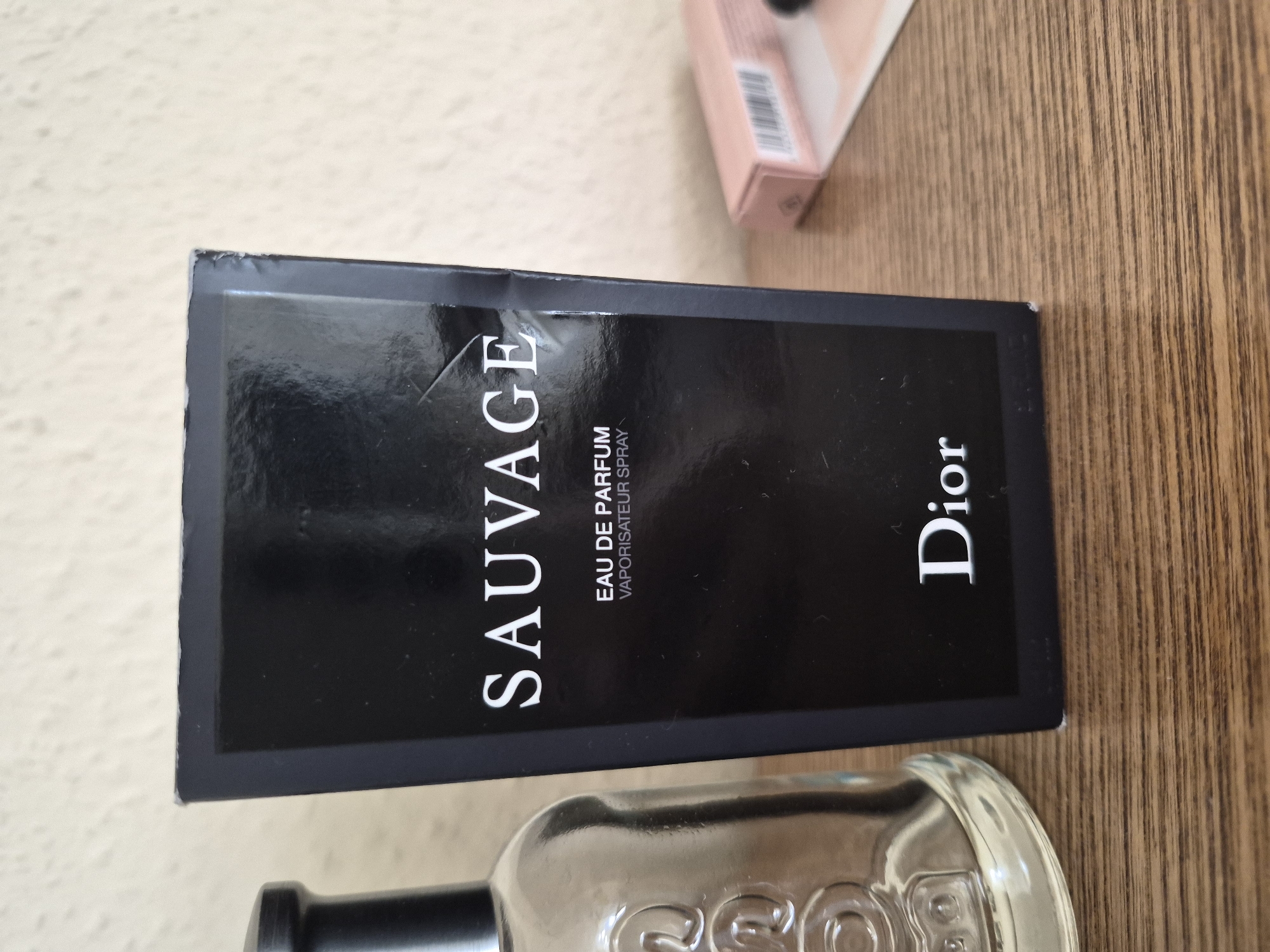 An upright shot shows a black box of Dior Sauvage Eau de Parfum Vaporisateur Spray next to a clear glass bottle on a wooden surface. The box is positioned in the center of the frame, with the bottle on its left. The background is a neutral, textured wall. The box is glossy black with white text. The text "SAUVAGE" is prominently displayed near the top of the box, followed by "EAU DE PARFUM" and "VAPORISATEUR SPRAY" in smaller text. The bottom of the box features the "Dior" logo in white. The glass bottle on the left is partially visible and appears to have embossed text on its surface. To the right of the box, a small, opened pink box with a barcode is visible. The lighting is soft, and the focus is primarily on the perfume box.