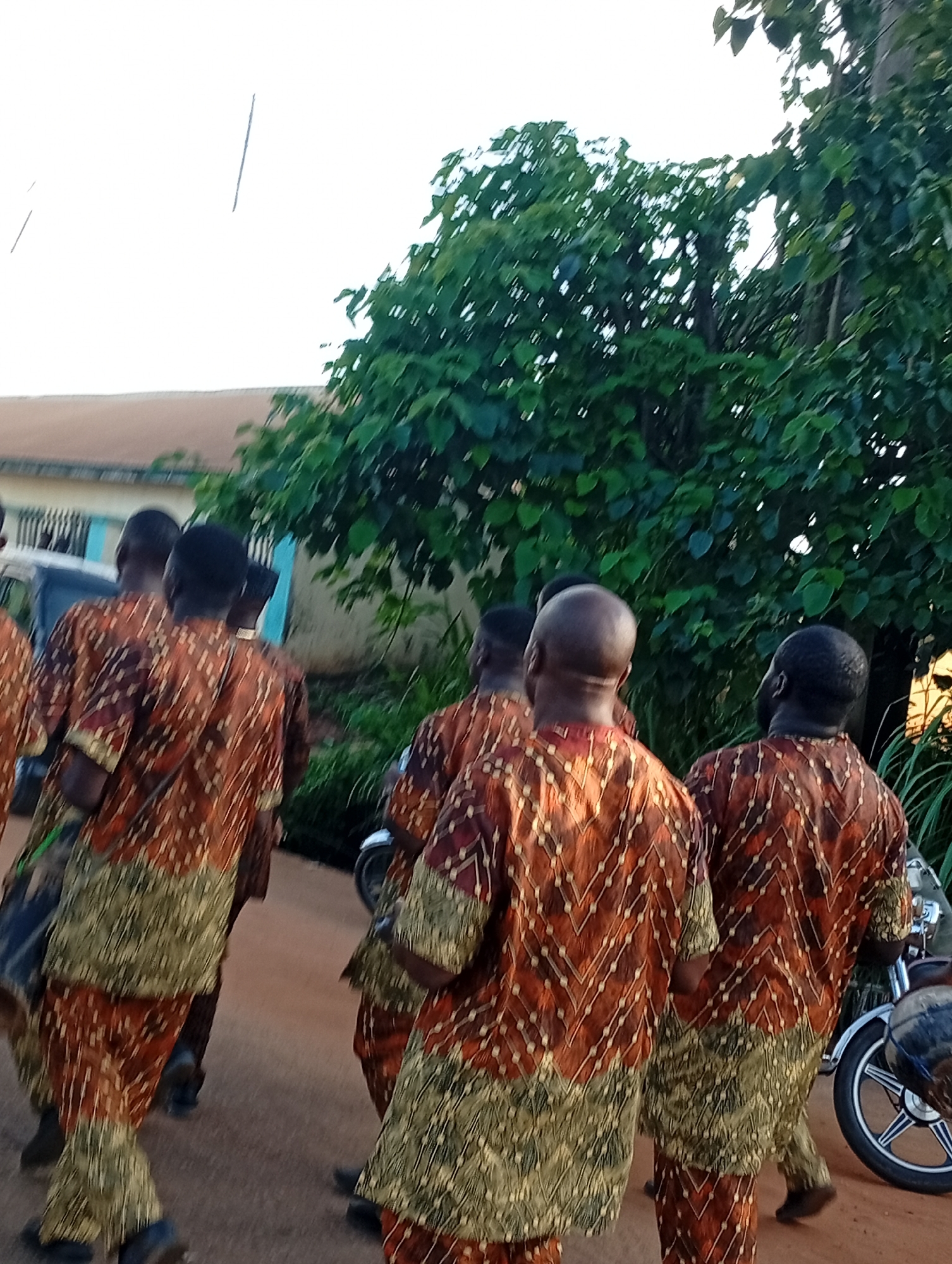 In Nsugbe, Nigeria, a group of men dressed in matching brown and gold patterned traditional attire are walking away from the camera on a dirt path. They appear to be in a procession or gathering. Behind them, a building with a light brown roof and light blue trim is visible, along with lush green trees and foliage. The sky is bright and overcast. A motorcycle is partially visible to the right of the frame. The scene suggests an outdoor cultural event or ceremony.