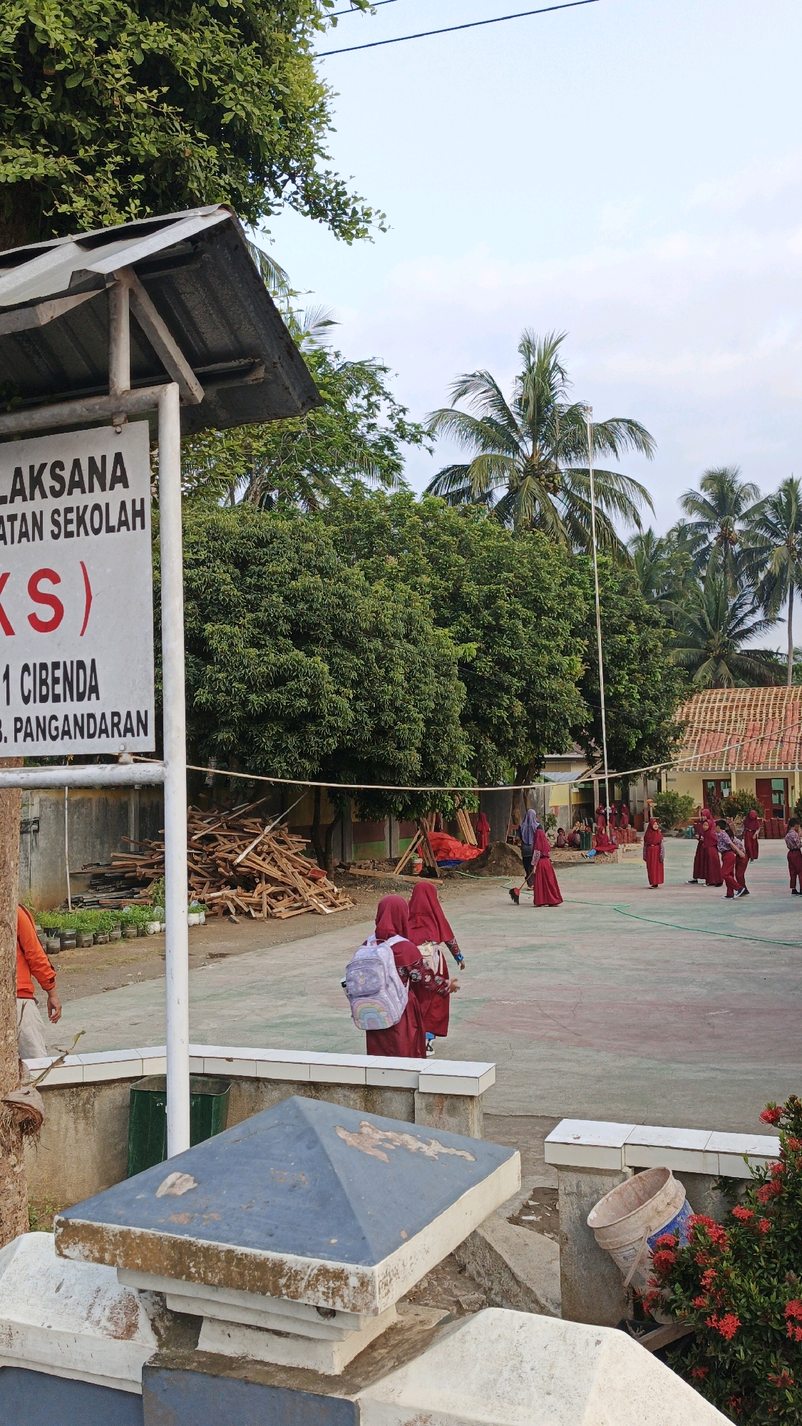 This image depicts an outdoor scene in Cibenda, Indonesia, likely a schoolyard based on the presence of students in uniform and a sign indicating school information. In the foreground, a signpost with a corrugated metal roof is visible, bearing text in Indonesian, including "PELAKSANA" and "ATAN SEKOLAH," suggesting it pertains to school operations. The sign also lists "CI BENDA" and "PANGANDARAN," confirming the location.
In the middle ground, several children, predominantly girls wearing maroon hijabs and uniforms, are seen on a paved court. Some are walking, while others appear to be engaged in activities. One girl is carrying a light-colored backpack with colorful cartoon images. In the background, lush green trees and palm trees are abundant, characteristic of a tropical environment. A building with a tiled roof is partially visible on the right side of the frame, further suggesting a school or institutional setting. Piles of wooden planks are stacked on the left side of the court, possibly indicating ongoing construction or maintenance. The sky is clear with some light clouds, suggesting daytime. The overall impression is of a lively school environment in a rural or semi-rural Indonesian setting.