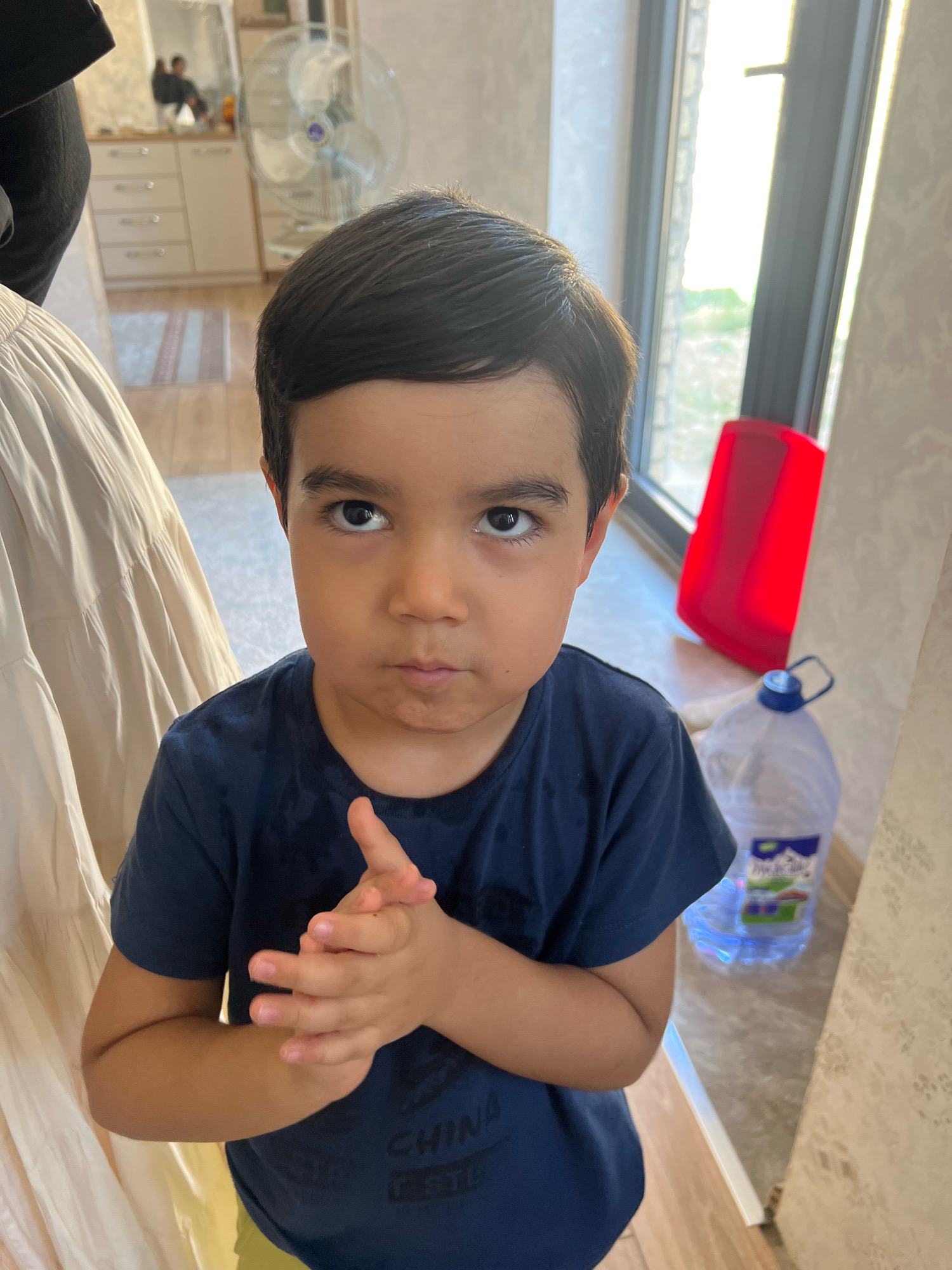 A young boy with dark hair and brown eyes is shown from the chest up, looking directly at the camera with a slightly furrowed brow and pursed lips. He is wearing a dark blue t-shirt with some text visible on it and yellow shorts. His hands are clasped together in front of his chest. The background is blurred but appears to be an indoor setting with a window, a large water bottle, a red object, and a fan. The lighting suggests it is daytime.