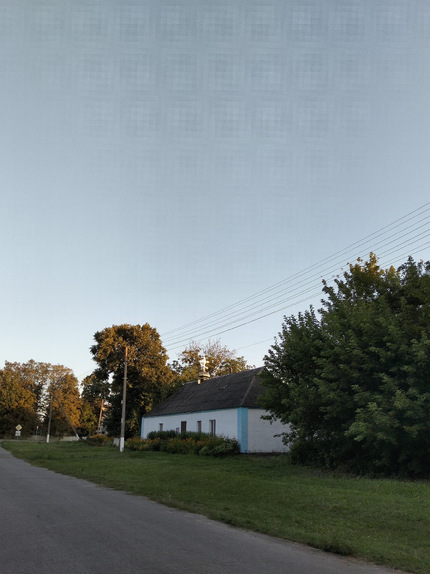 The image depicts a rural scene in Kliushnykivka, Ukraine. In the foreground, a paved road curves to the left, bordered by a grassy verge. To the right of the road, a white building with a blue stripe under the roof is situated. The building has a dark, textured roof and several windows. In front of the building, a row of flowering bushes adds color to the scene.
Behind the building and to the right, a dense canopy of green trees dominates the mid-ground. To the left of the building and further back, several tall trees with golden-brown foliage are visible, suggesting it might be autumn. A utility pole with wires stretching across the frame stands near these trees. Another utility pole is positioned closer to the building, with white markings at its base. In the far left background, a yellow diamond-shaped road sign is visible on a post. The sky above is a clear, pale blue with a subtle grid pattern overlaid, possibly an artifact of image processing. No people or animals are present in the image.