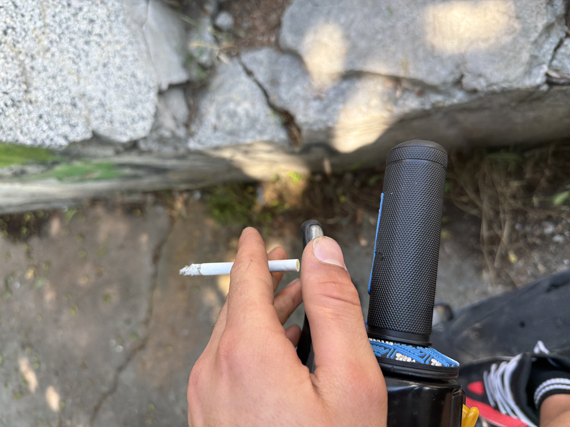 The image presents a first-person, downward-angled view of a person's left hand holding a lit cigarette, positioned above the handlebar of what appears to be a bicycle or similar vehicle. The scene is outdoors in Basarbovo, Bulgaria, under bright daylight.
The left hand, exhibiting fair skin and short fingernails, holds a white cigarette vertically between the index and middle fingers. The cigarette's tip displays a substantial amount of grey ash, indicating it has been burning. The filter end of the cigarette is a light yellow-orange color. The hand is resting over a black, textured rubber grip of a handlebar, which features a diamond or criss-cross pattern. Adjacent to the grip is a blue plastic ring with a stylized pattern, followed by a black plastic housing, likely part of a brake or gear mechanism. A yellow lever, possibly a brake lever, is partially visible beneath the black housing.
In the lower-left corner, a portion of the person's left foot, wearing a black and red athletic shoe with white laces and white side stripes, is visible on a dark surface, suggesting the pedal or footrest area of the vehicle.
The background consists of an uneven, cracked concrete or asphalt ground, scattered with small green weeds, dried leaves, and debris. To the right, a rough-textured, light grey concrete or stone wall rises, showing irregular breaks and some faint green discoloration, possibly moss or graffiti. Strong sunlight illuminates the scene, casting defined shadows, notably from the hand and the wall.