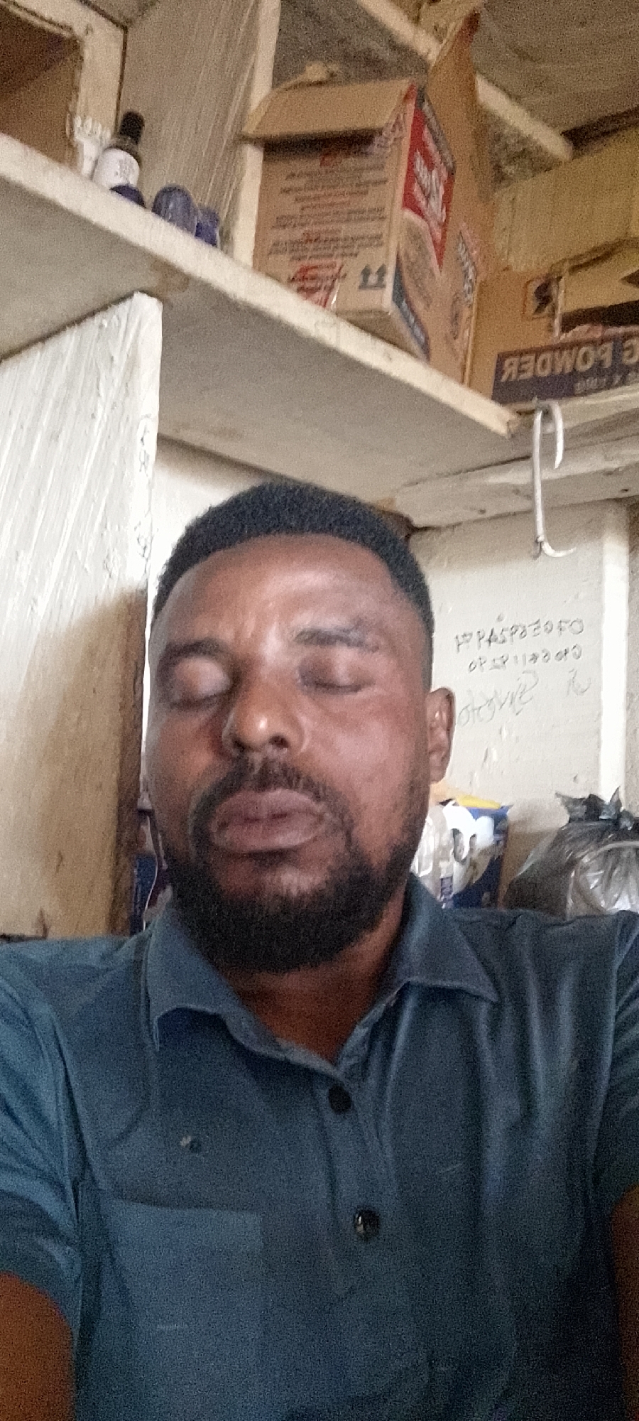 A medium shot captures a Black male with dark skin, short black hair, and a beard, looking upwards with his eyes closed. He is wearing a blue collared shirt. Behind him, shelves are visible, holding various items including boxes, bottles, and what appears to be a roll of plastic wrap. The environment suggests an indoor setting, possibly a shop or storage area, with a white wall to the left and a lighter-colored wall behind him. The lighting is somewhat dim, casting shadows. The overall impression is of a candid, informal moment. The image location is identified as Durumi, Nigeria.