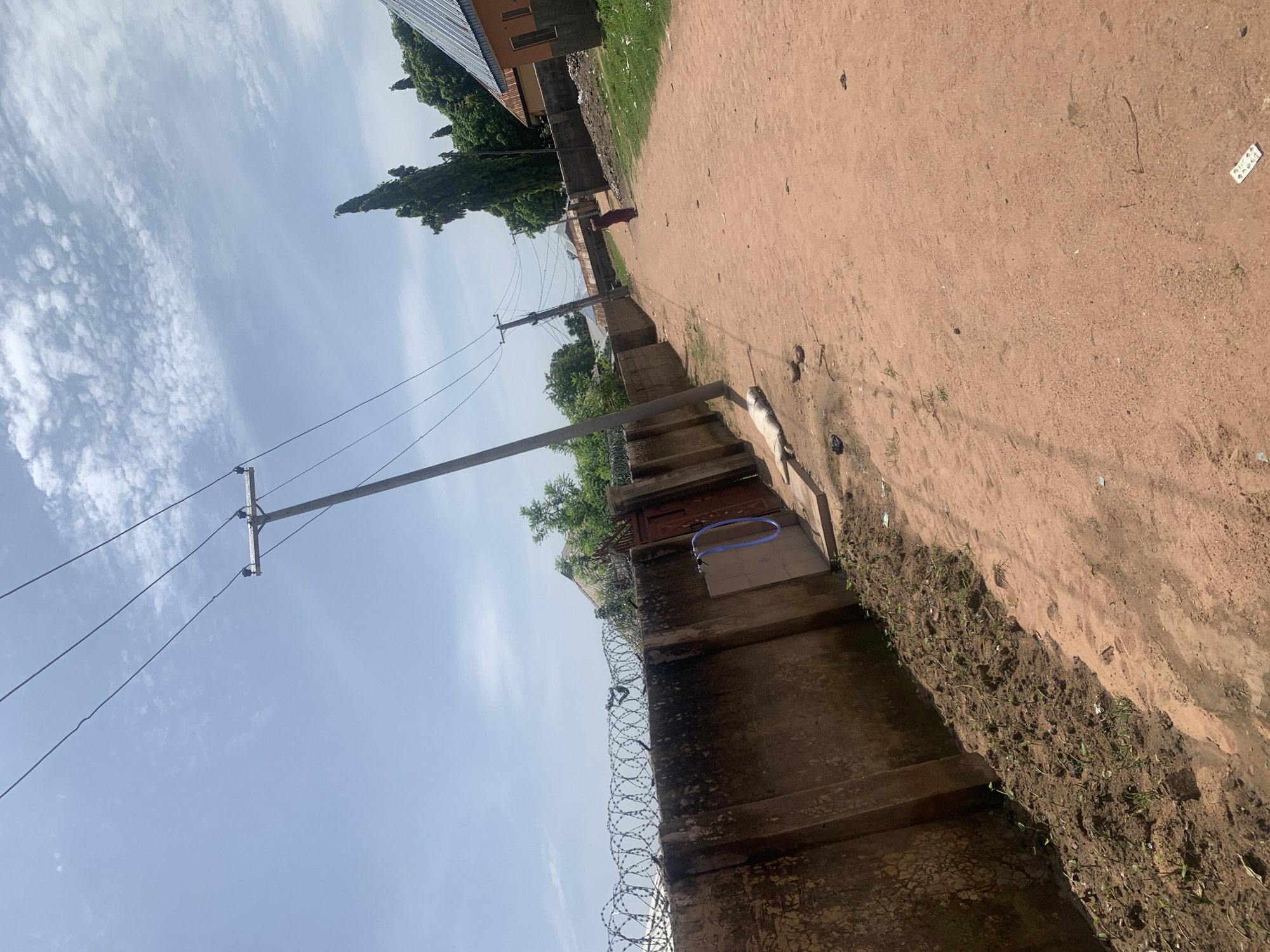 The image depicts a dirt road in Jauro Yinu, Nigeria. On the left, a concrete wall topped with barbed wire runs along the property line. Several electrical poles with wires are visible, with one pole leaning significantly. A person wearing a dark red garment is walking down the road in the distance on the right. Houses with corrugated metal roofs are situated on either side of the road. The sky is partly cloudy. The overall scene is a clear day in a rural or semi-rural Nigerian setting.