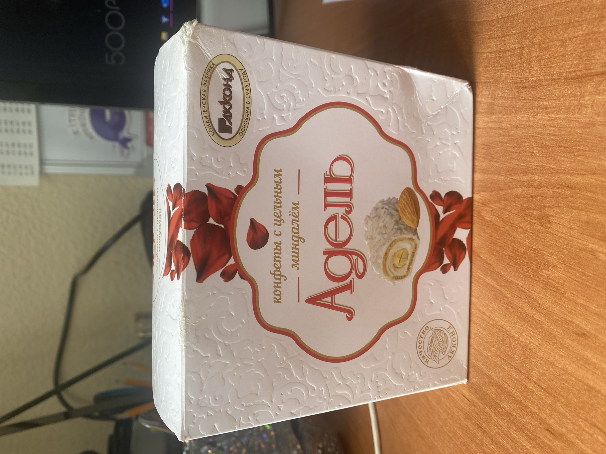 A white cardboard box of chocolates sits on a wooden surface. The box has red and gold floral embellishments and text in Russian. The text identifies the chocolates as "Адель" from the confectionary factory "Акконд" founded in 1943. A circular emblem on the box reads "качество Акконд". The background shows a desk with office supplies and a computer monitor. The scene is indoors.