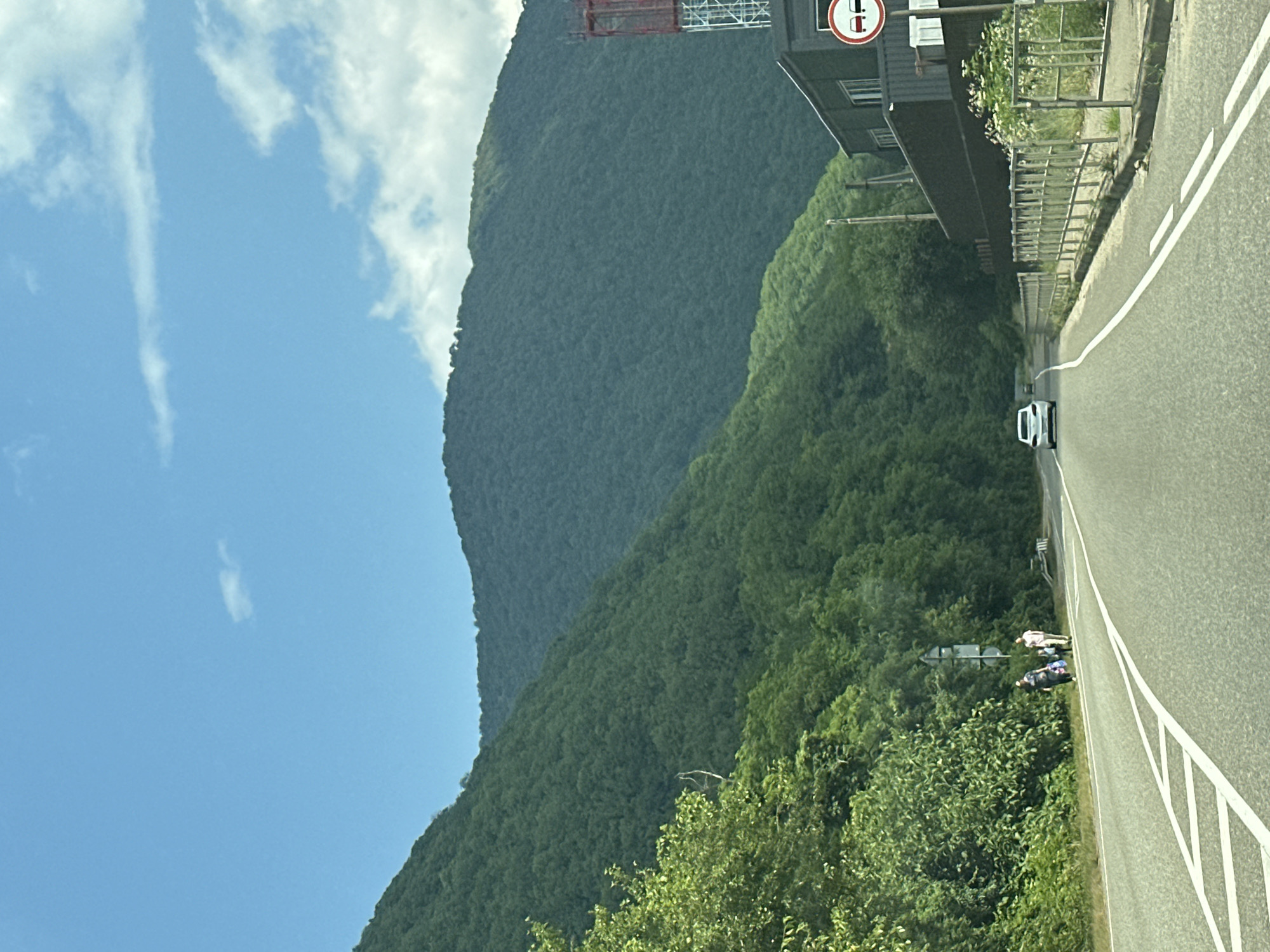 The image depicts a scenic drive with lush green mountains framing a two-lane asphalt road. A white car is driving away from the camera, down the road. On the left side of the road, dense vegetation covers the mountainside, with some trees reaching towards the sky. A few people are walking on the shoulder of the road on the left, carrying bags. On the right side of the road, a dark-colored building with a tall red and white structure behind it is visible. A road sign indicating "no overtaking" is mounted on a pole in front of the building. The sky is a clear blue with scattered white clouds. The scene appears to be a highway passing through a mountainous, forested area, possibly on the outskirts of Tuapse, Russia, given the context provided.