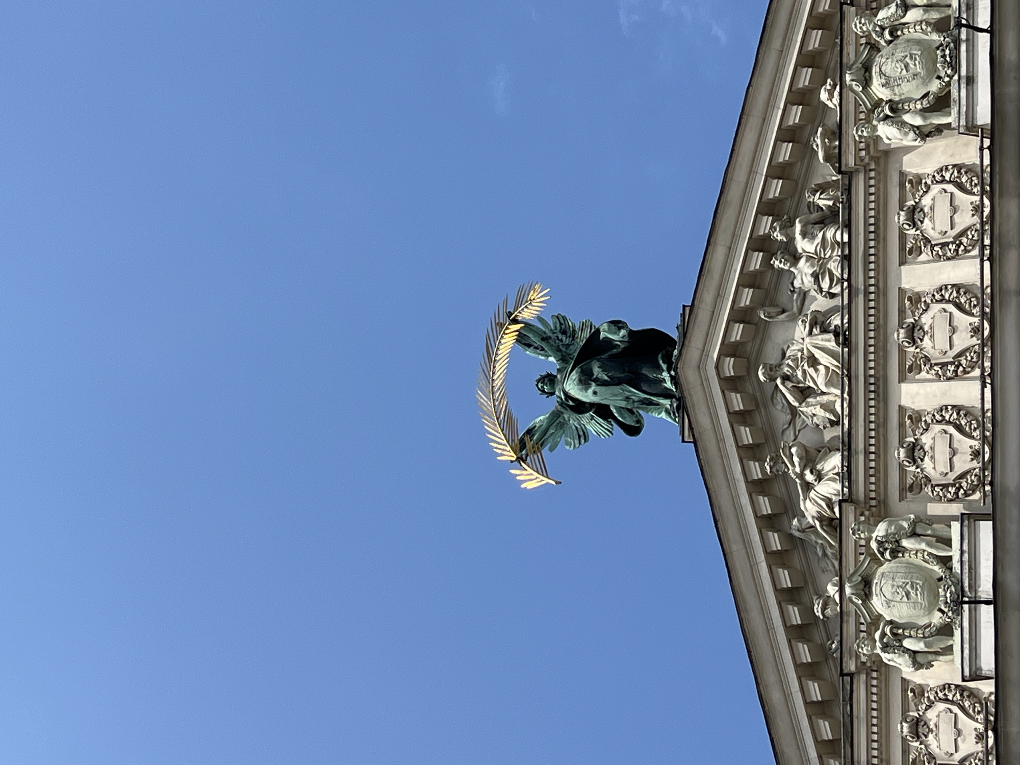The image captures the upper portion of the facade of the Lviv Opera House, a prominent landmark in Lviv, Ukraine. A bronze statue of an angel holding a golden laurel wreath stands atop the building's roof. Below the angel, the roofline is adorned with a series of sculpted figures and decorative elements, typical of Renaissance architecture. The sky is a clear, bright blue, with a few wispy clouds visible. The overall impression is one of grandeur and historical significance, showcasing the architectural beauty of the opera house against a clear sky.