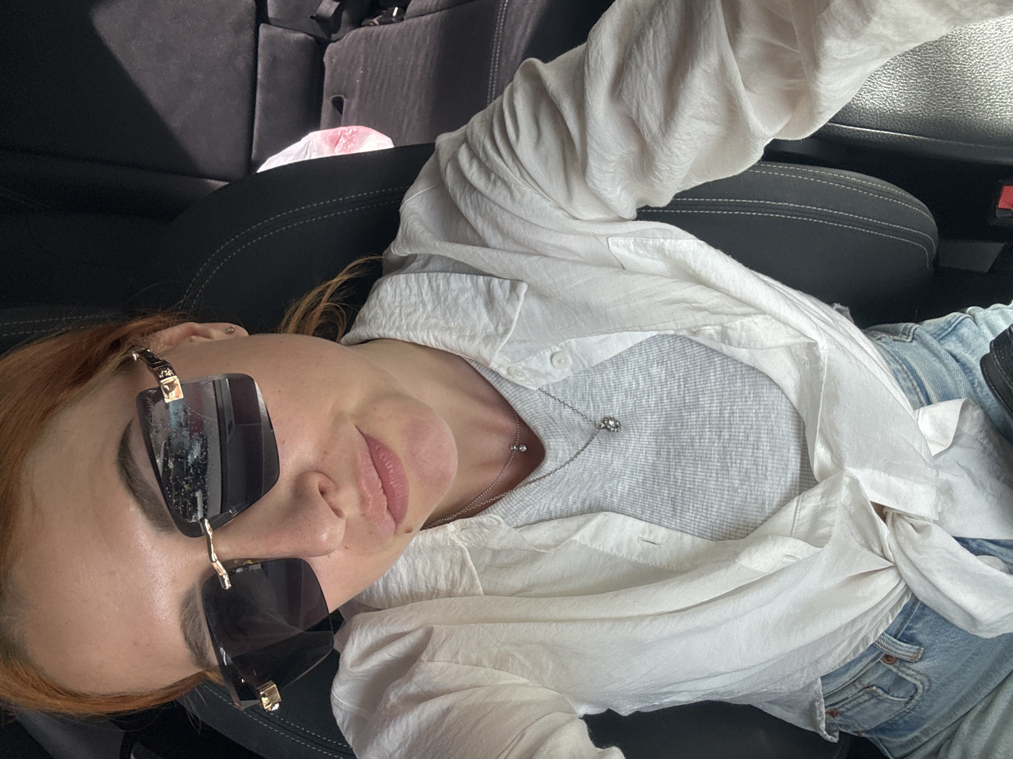 The image captures a person reclining casually in the passenger seat of a car, positioned diagonally across the frame. The subject has fair skin and reddish-brown hair, partially obscured by large, dark-tinted rectangular sunglasses with prominent gold-colored frames. Their lips are slightly parted in a relaxed manner, suggesting a calm or subtle smile.
They are dressed in a casual ensemble featuring a white, crinkled long-sleeved button-up shirt worn open over a gray ribbed tank top. A delicate silver-colored chain necklace with a small pendant, composed of two closely set clear stones or charms, rests on their chest. A small, silver-colored stud earring is visible in their left ear. Light blue denim jeans are partially visible on the right side of the frame, peeking out from under the white shirt.
The setting is the interior of a vehicle, identifiable by the dark gray or black car seats and upholstery visible in the background and surrounding the person. A hint of an armrest or door panel is also discernible. Natural light illuminates the scene, suggesting it is daytime. A blurry, light pink object, possibly a plastic bag, can be seen in the distant background. The overall mood is relaxed and unposed. This photo was taken in Budva, Montenegro. There is no visible text in the image.