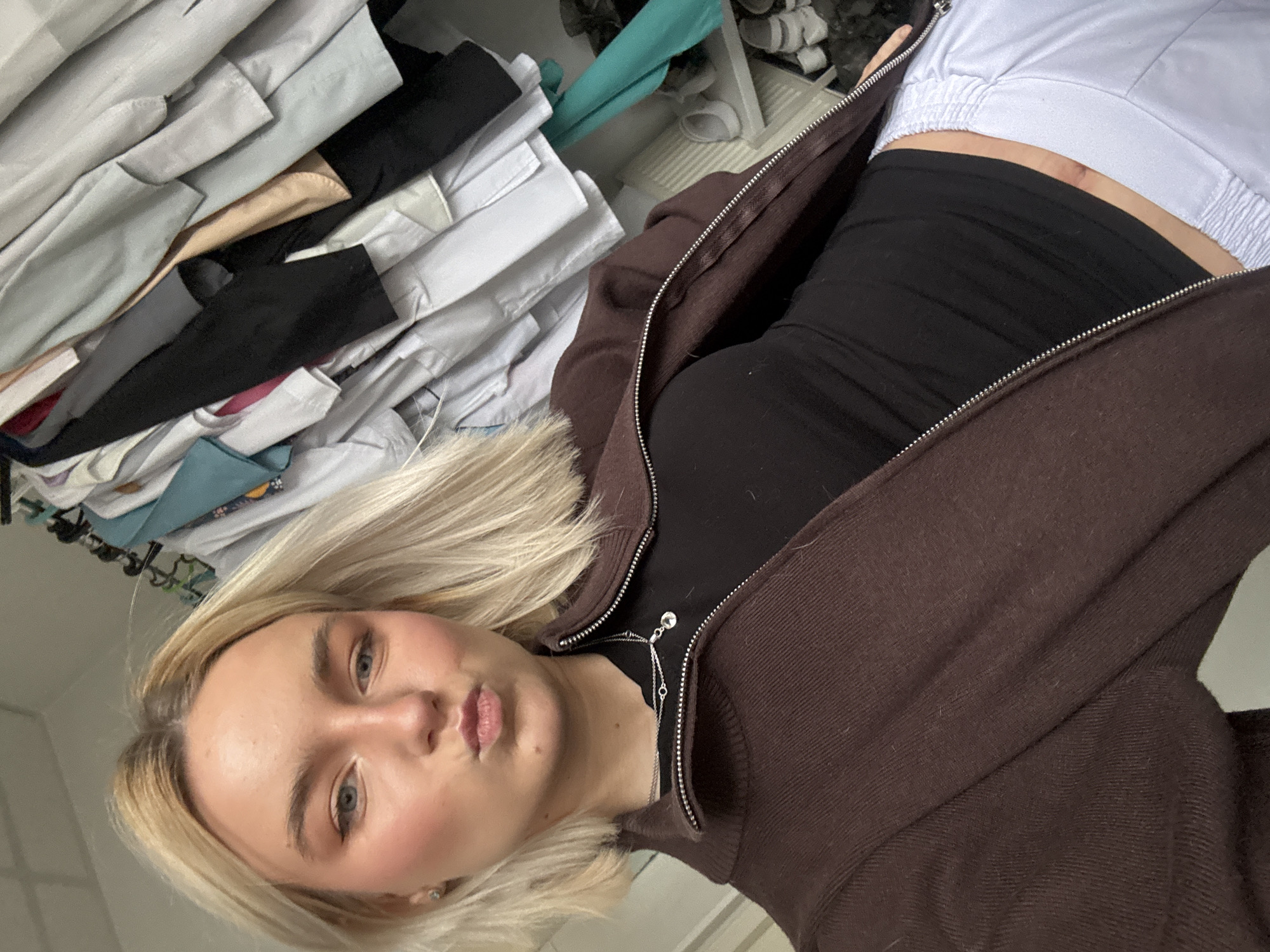 The image appears to be a selfie taken from a slightly angled, upward perspective, showing a young woman with fair skin and blonde hair. She is wearing a black top and a brown zippered jacket, with white bottoms visible in the upper right corner of the frame. She is also wearing a necklace. The background is cluttered with various items, including what appears to be a stack of clothing and shoes. The location is not discernible from the image and cannot be attributed to Saint Petersburg, Russia, or any other specific city.