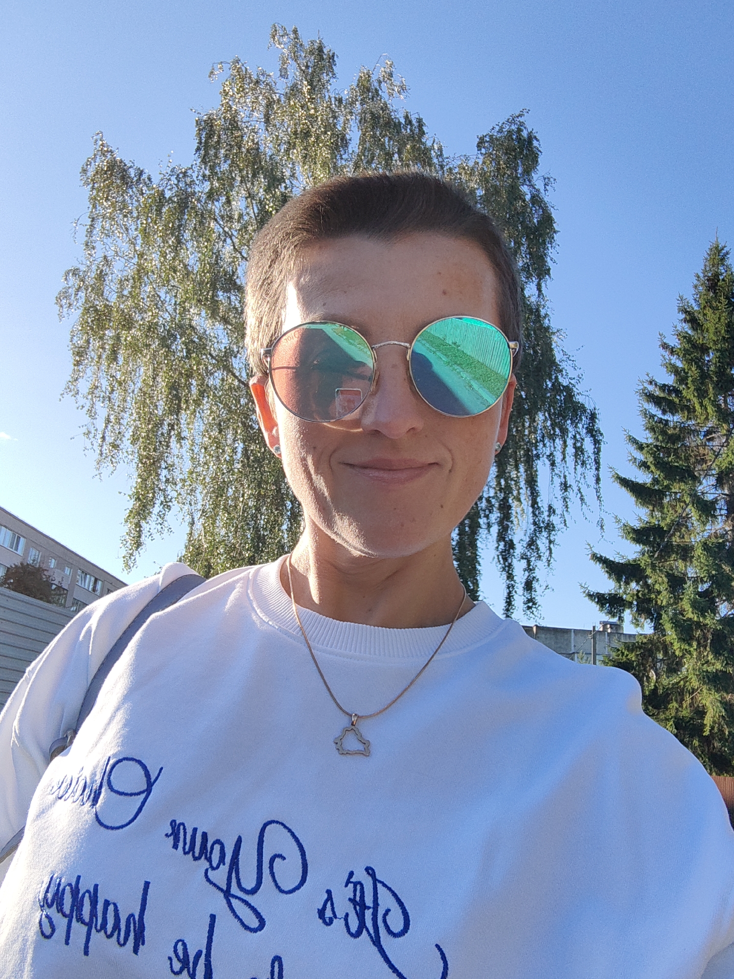 A person is taking a selfie outdoors during the day. The person is wearing a white sweatshirt with blue cursive writing and a gold necklace with a cloud-shaped pendant. They are also wearing round sunglasses with iridescent lenses. Behind the person, there are trees and a building. The sky is blue and clear. This scene is located in the city of Fanipal, Belarus.