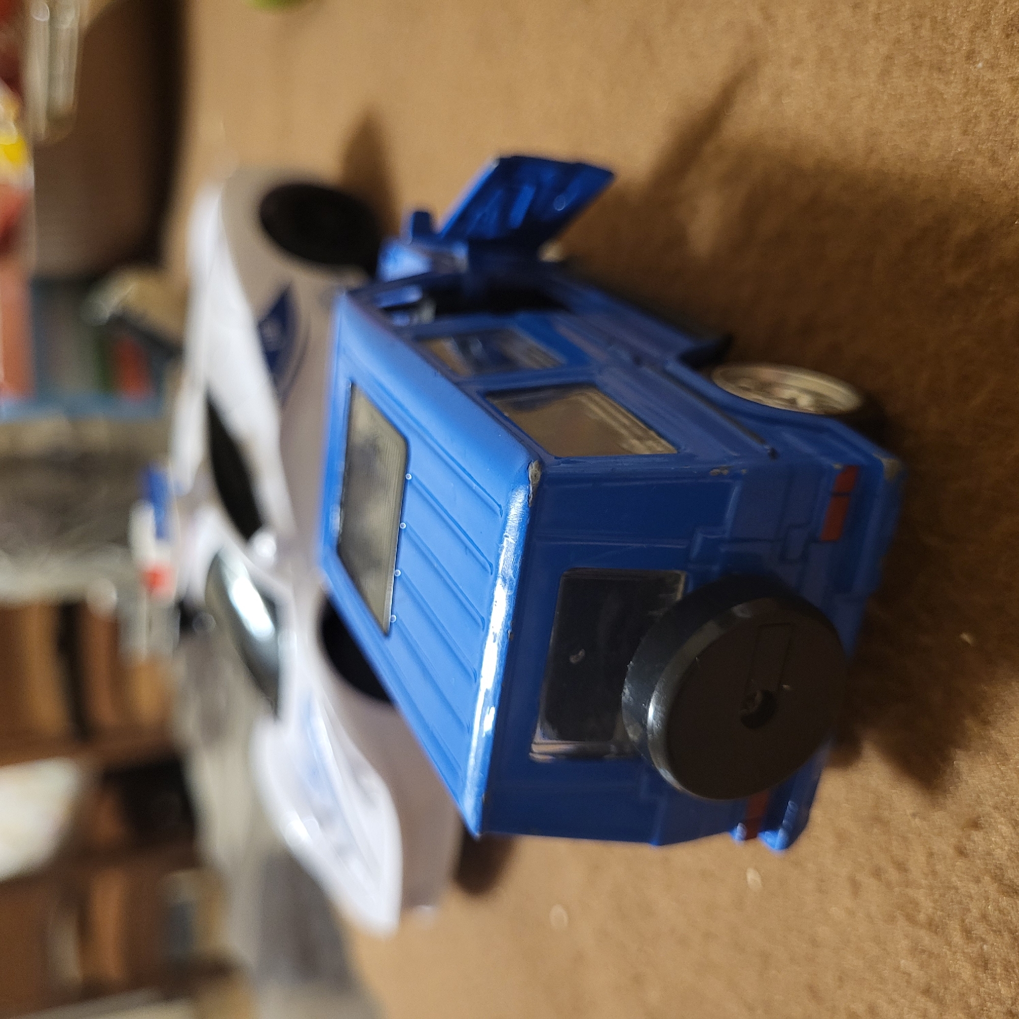 A partially focused overhead shot shows two toy cars on a tan surface. One car is blue, a boxy, off-road-style vehicle with the hood propped open. A spare tire is mounted on the back. The second car is white and more aerodynamic. The background is blurred, suggesting a room interior. There is no indication in the image that the location is Yerevan, Armenia.