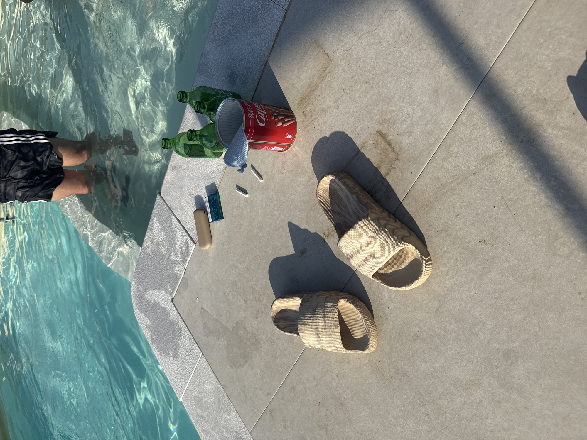 The image shows a close-up view of a swimming pool area at the city of Potos, Greece. In the foreground, a person wearing black shorts with white stripes is standing in the shallow end of a turquoise swimming pool. The pool edge, made of light-colored textured tiles, is visible. On the tiled deck next to the pool are two light-brown, ribbed slide sandals. To the right of the sandals, a red cylindrical can with a silver lid, labeled "Capri," is placed on the tiles. Inside the can, chocolate biscuit sticks are visible. Next to the can are three green glass bottles. Further to the left, a small rectangular beige object, possibly a case, and a pack of cigarettes with "TEREA" printed on it, along with two white sticks, possibly cigarette sticks, are lying on the tiles. The sun is casting long shadows across the scene, indicating it is likely late afternoon or early morning.
