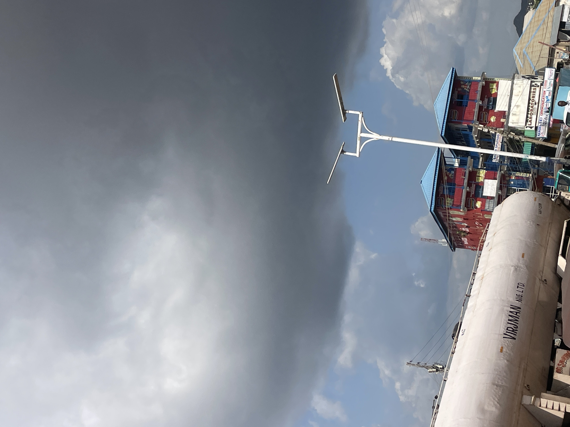 The image depicts an outdoor scene in Rafin Par, Nigeria, dominated by a dramatic sky. Heavy, dark storm clouds fill the upper portion of the frame, contrasting with patches of brighter, blue sky and fluffy white clouds below. In the foreground, a large white tanker truck with the name "VIRJMAN NIG. LTD." is visible, partially obscuring the view of the buildings behind it. To the right of the tanker are several multi-story buildings with red and blue facades, featuring balconies and signs for businesses, including "LABORATORIES." A modern streetlamp with two white light fixtures stands prominently in the middle ground. The overall impression is a bustling urban environment under an imposing sky, suggesting an impending storm.