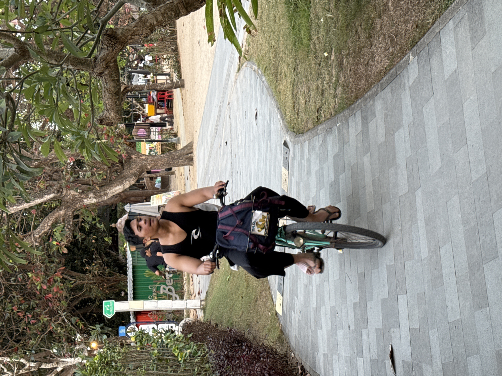 A young adult male with dark hair is riding a light green bicycle on a paved pathway in Denpasar, Indonesia. He is wearing a black tank top, black pants, and flip-flops. A dark backpack with red straps is secured to the front of the bicycle. The pathway is lined with green grass and trees. In the background, there are buildings, other people, and signs. The overall setting appears to be an outdoor public space, possibly a park or promenade.