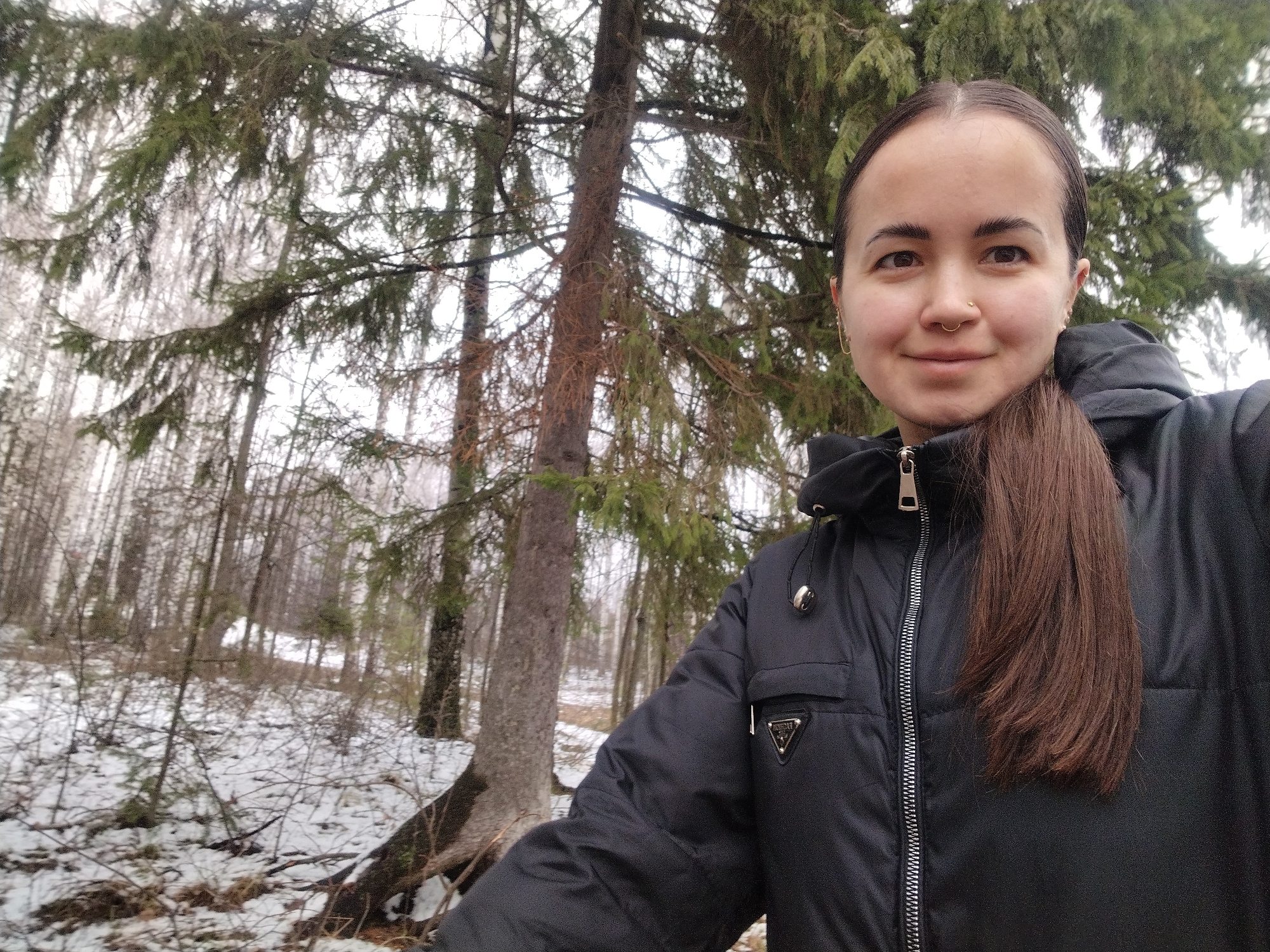 A woman stands in a snowy forest, likely in the vicinity of Noviy, Russia. She is wearing a black, hooded winter jacket with a visible zipper and a triangular logo on the chest pocket. Her brown hair is pulled back into a ponytail. She faces the camera with a slight smile. Behind her, the forest shows scattered snow cover and a mix of deciduous and evergreen trees. The scene appears overcast with flat lighting.