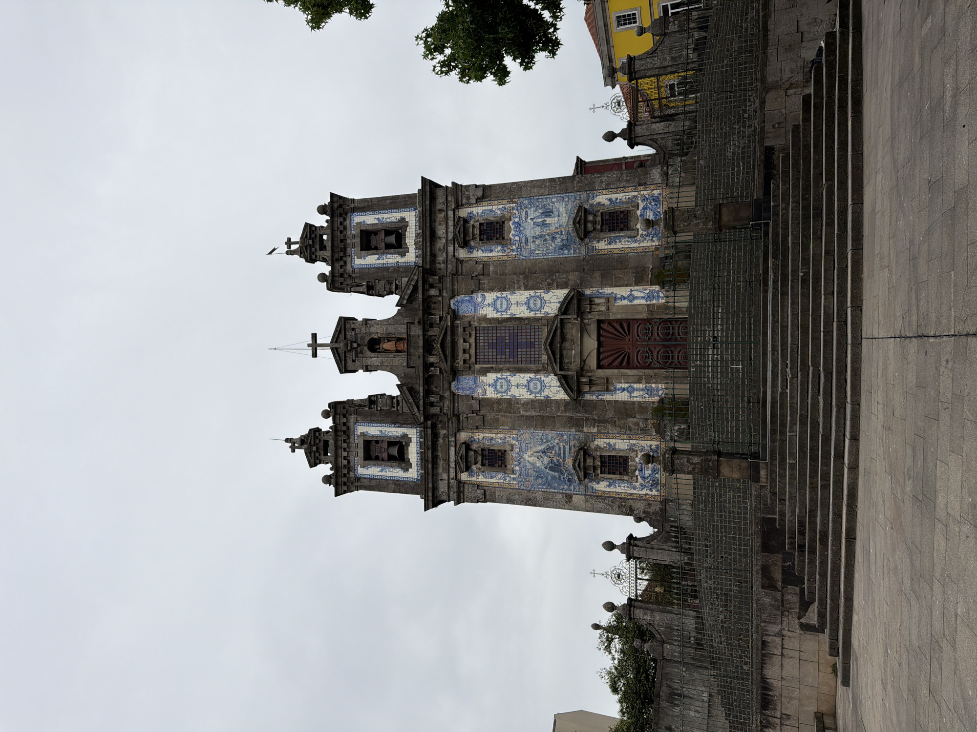 The image shows the Igreja do Carmo in Porto, Portugal. It is a baroque church known for its facade decorated with azulejo tiles, which depict religious scenes. The church has two towers and a large staircase leading up to the entrance. The sky is overcast, and the lighting is diffused. The church is surrounded by a stone wall and a metal fence. There are trees and a yellow building visible in the background.