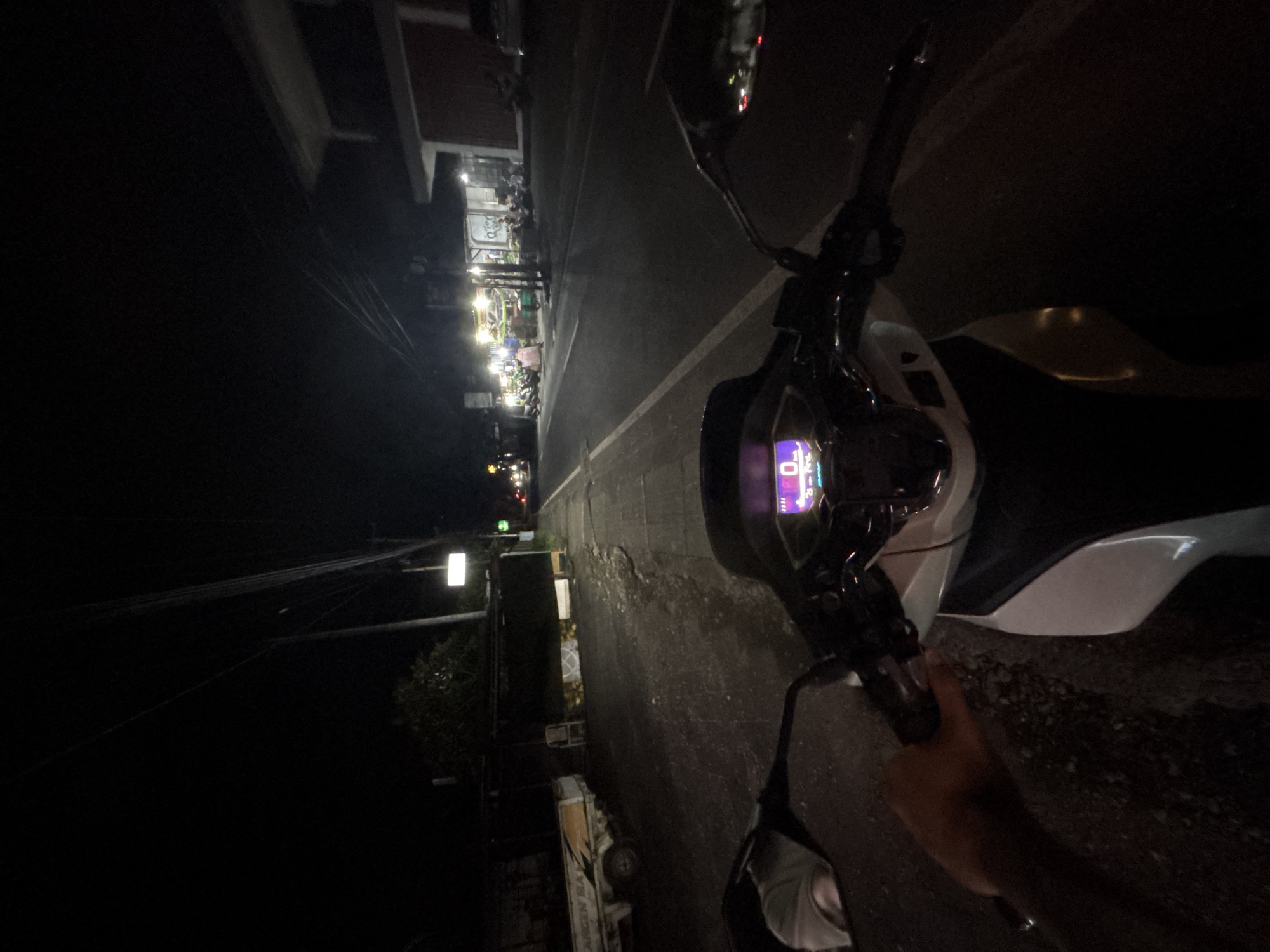 This media file captures a nighttime street scene from the first-person perspective of someone on a motorcycle or scooter in Kuta Selatan, Indonesia.
In the foreground, the handlebars, a rearview mirror, and parts of a white and black motorcycle are prominently visible. The digital instrument cluster is lit up with a purple glow, displaying "0" or "00." A human hand is seen resting on the right handlebar.
The scene unfolds along a dark street, likely asphalt or concrete, with a white line delineating its right edge. To the left, a row of small commercial establishments or shops are brightly illuminated by their internal lights, casting a glow onto the road. Several people are visible in front of these shops, some appearing to be patrons or staff, and numerous motorcycles or scooters are parked along the sidewalk. Further down the street on the left, a light-colored truck or van is partially visible, bearing the distinct text "LANGSUNG BAI" on its side.
The sky is dark, indicative of nighttime, and overhead power lines or communication cables crisscross the upper portion of the frame. The overall impression is that of a bustling local street in the evening, with ongoing commercial and social activity.