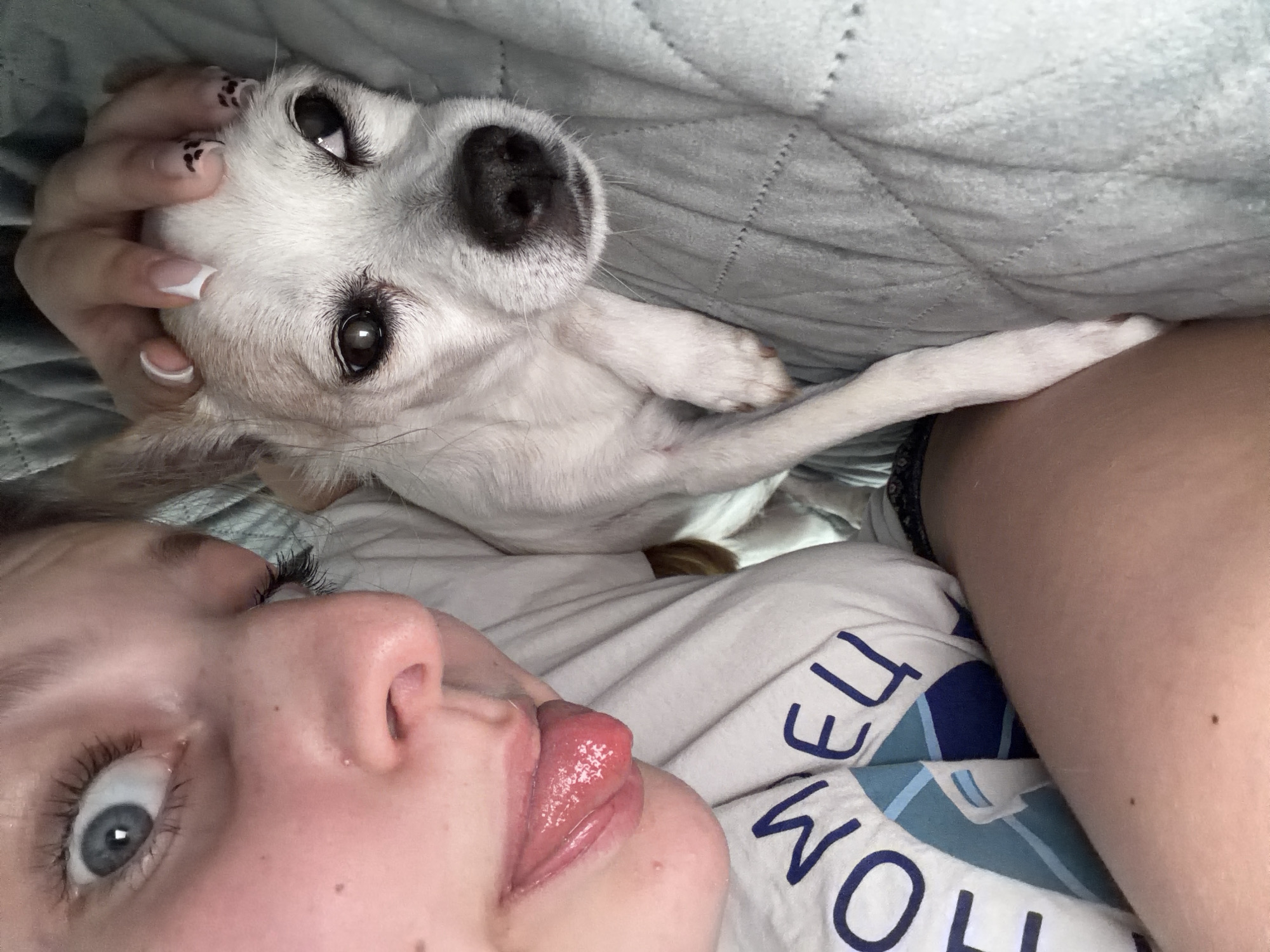 The image shows a close-up of a young person with their face tilted downwards, sticking out their tongue and looking at the camera. They are lying down on a light blue quilted blanket, with a small white dog resting beside them. The person is wearing a white t-shirt with blue lettering. The dog is white with light brown ears and a black nose. The person's left arm is visible on the left side of the frame, showing two small moles. The setting is indoors, likely a bedroom, with no specific identifying features of Burgas, Bulgaria visible.