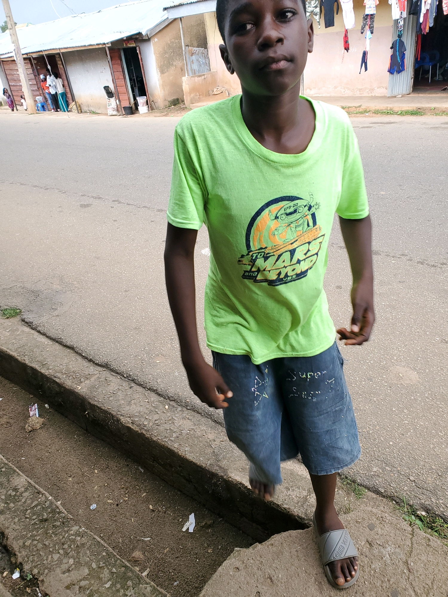 A young Black boy walks down a dusty road in Jos, Nigeria, wearing a bright green t-shirt and denim shorts. He is looking forward with a neutral expression. In the background, a row of small shops with corrugated metal roofs are visible. Clothes are hung out to dry in front of one of the shops. The ground is a mix of asphalt and dirt, with a drainage ditch running along the side of the road. There are a few pieces of trash scattered around. The boy is wearing gray sandals.