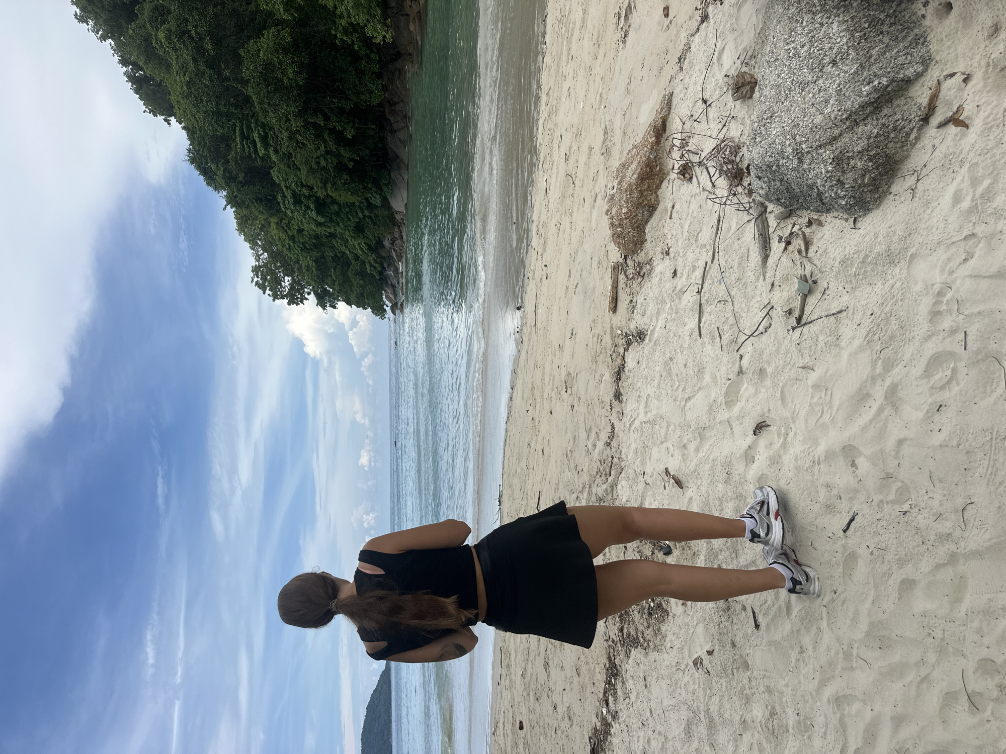 A woman stands on a sandy beach, looking out at a tranquil turquoise bay with lush green hills rising on the right. The sky is a mix of blue and white clouds. The woman, viewed from behind, wears a black crop top and a black skirt, with athletic shoes on her feet. The beach is strewn with small pieces of driftwood and rocks. In the distance, a small boat can be seen on the water. The scene suggests a peaceful moment in a tropical setting, possibly Karon, Thailand, given the distinctive landscape and beach characteristics.