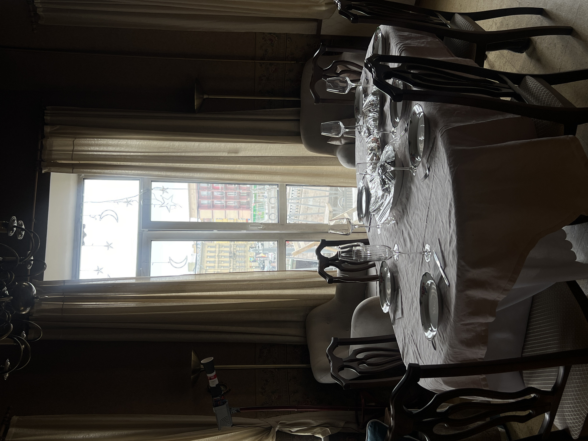 The image depicts an interior scene, likely a dining room, in Saint Petersburg, Russia. A long table is prominently featured, set with plates, silverware, and glassware. Several dark-colored chairs surround the table. A large window with decorative celestial shapes is visible in the background, offering a view of a city street, identifiable by the architecture as Saint Petersburg. Drapes hang on either side of the window, adding to the ambiance of the room. A chandelier is partially visible in the lower left corner. The room is somewhat dimly lit.