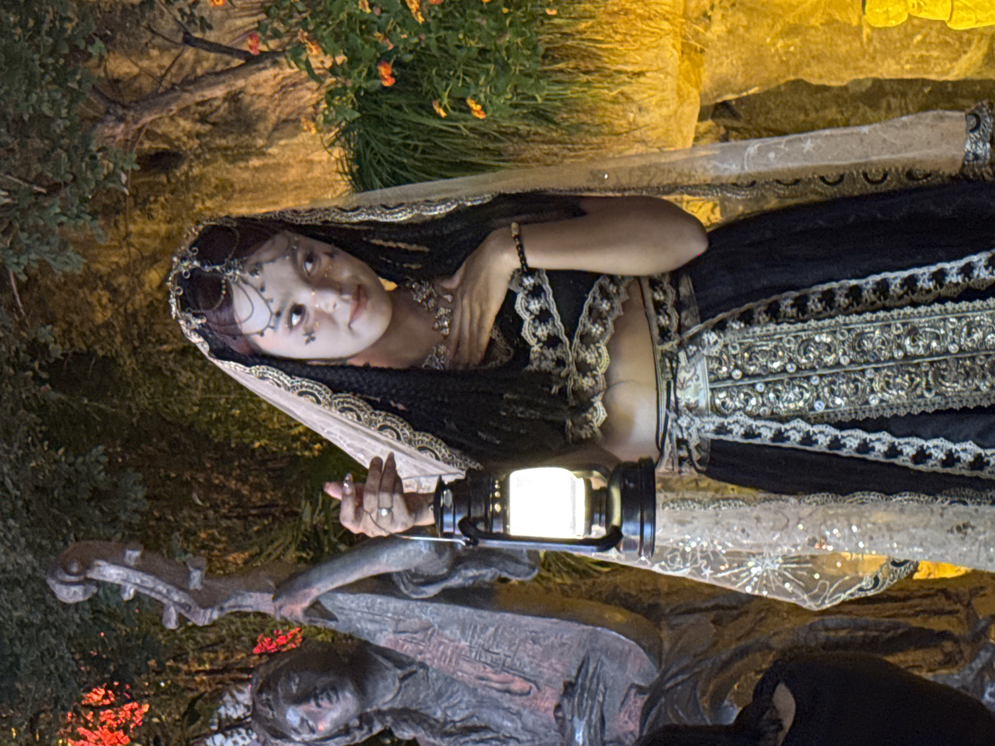 In Yuzhong District, China, a young woman dressed in ornate, traditional clothing stands in an outdoor setting at dusk. She wears a jeweled headpiece, a sheer veil with lace trim, a black top with intricate silver embellishments, and a long, flowing skirt also adorned with silver embroidery. Her makeup is dramatic, with elaborate markings around her eyes. In her right hand, she holds a lit oil lantern, its glow illuminating her face and the surrounding scene. To her left, a weathered, grey statue of a person playing a stringed instrument stands as a backdrop, its form partially obscured by foliage. The background consists of lush greenery and trees, with warm, artificial lighting casting a golden hue on the environment, suggesting a theatrical or decorative display.
