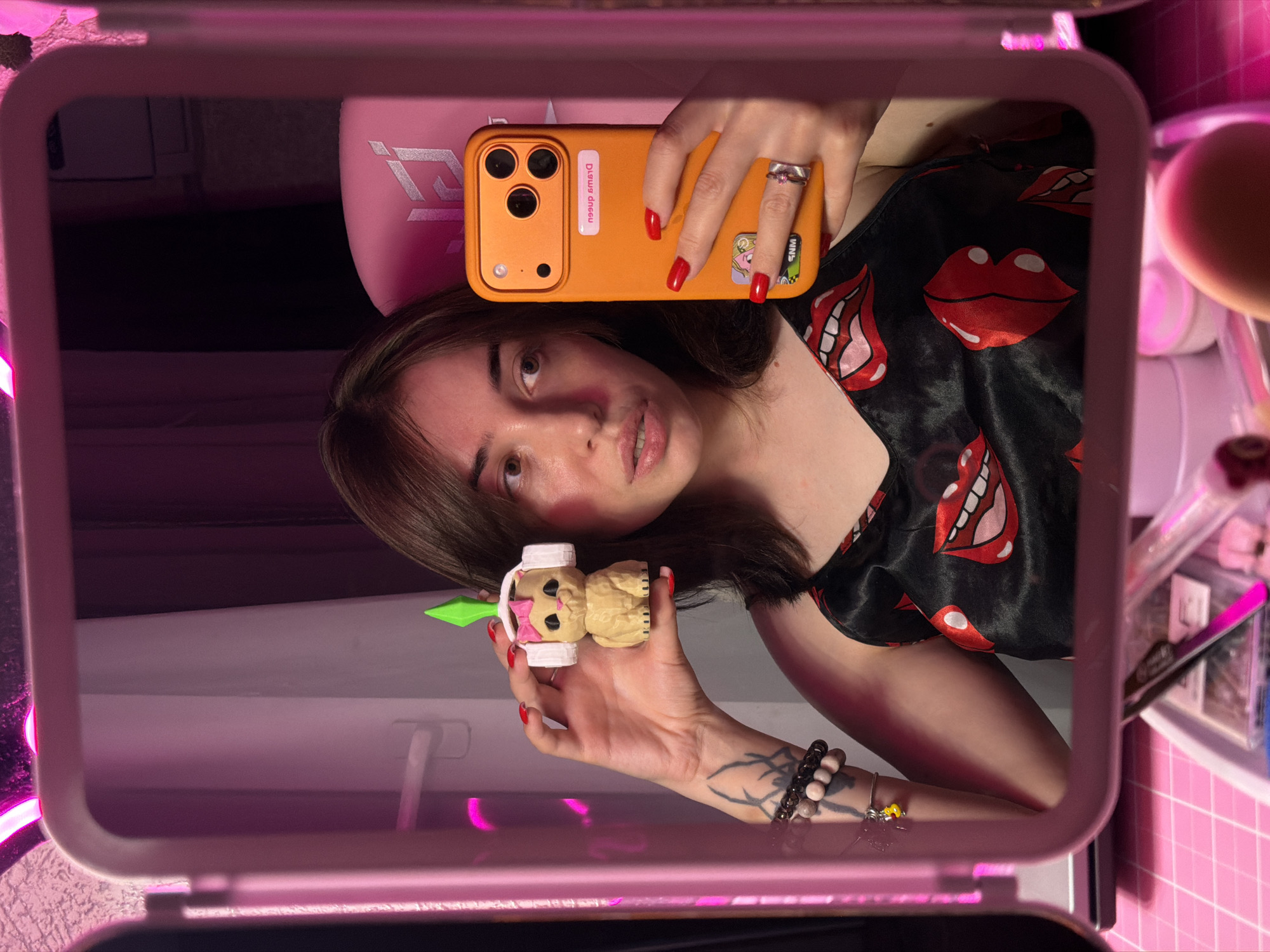 In a dimly lit room, a young woman with brown hair and fair skin takes a selfie in a mirror. She is wearing a black satin slip dress with a red lips pattern. In her right hand, she holds an orange smartphone, and in her left hand, she holds a small figurine of a cat wearing headphones and a green diamond on its head. Her nails are painted red, and she wears a silver ring on her right ring finger and a bracelet on her left wrist. The reflection also shows a pink gaming chair in the background and a vanity table with various cosmetic items. The overall scene is set against a backdrop of pink lighting, suggesting a personal and vibrant atmosphere. The image is taken from a frontal, slightly high-angle perspective, capturing the woman's upper body and the details of her reflection.
