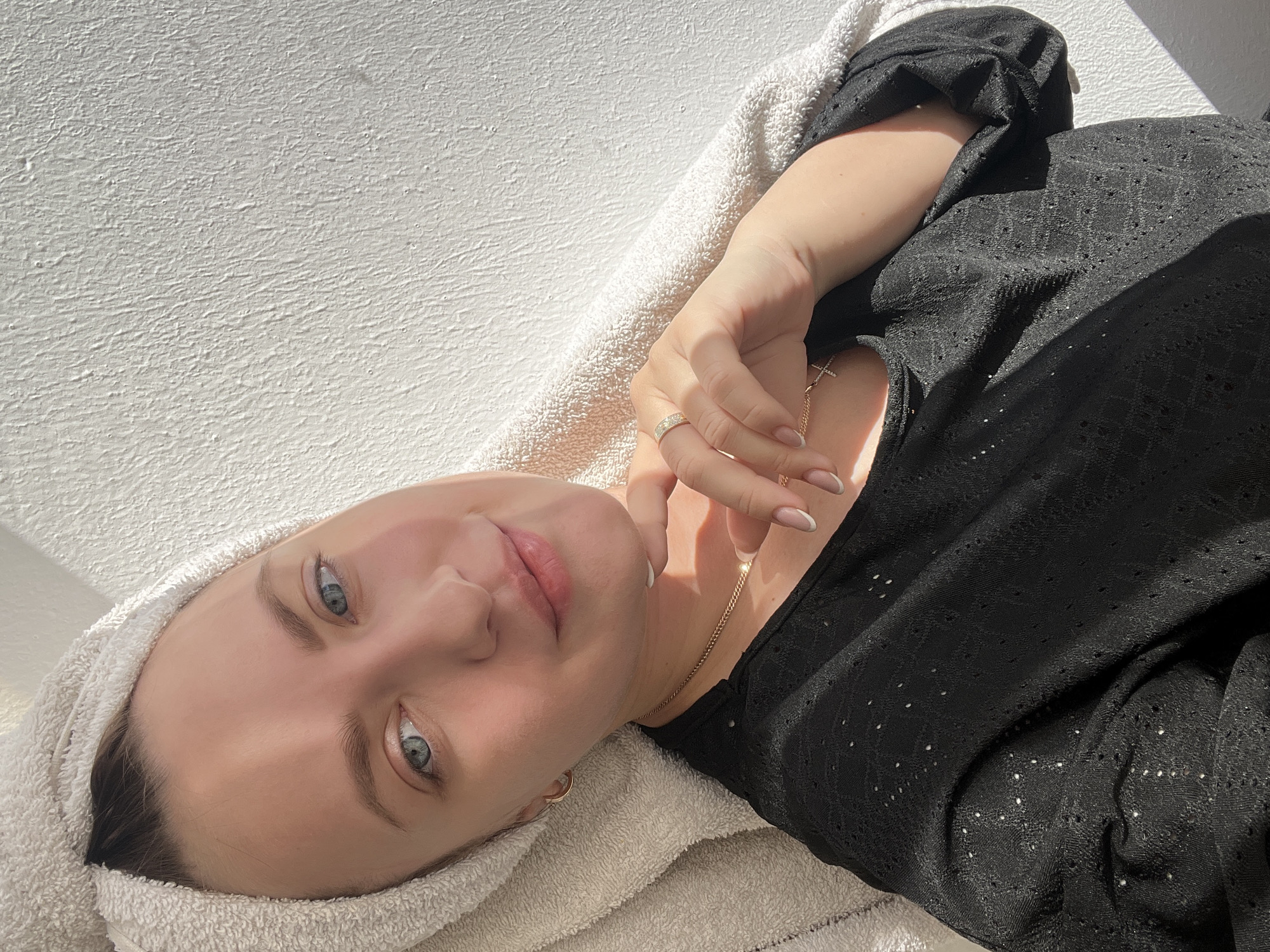 A woman lies on a light-colored towel in a well-lit space. She has light skin, blue eyes, and her hair is wrapped in the towel. She is wearing a black top and a ring on her finger. A gold necklace is visible around her neck. The location is uncertain and not identifiable as Марковский, Russia, from the visual information provided.