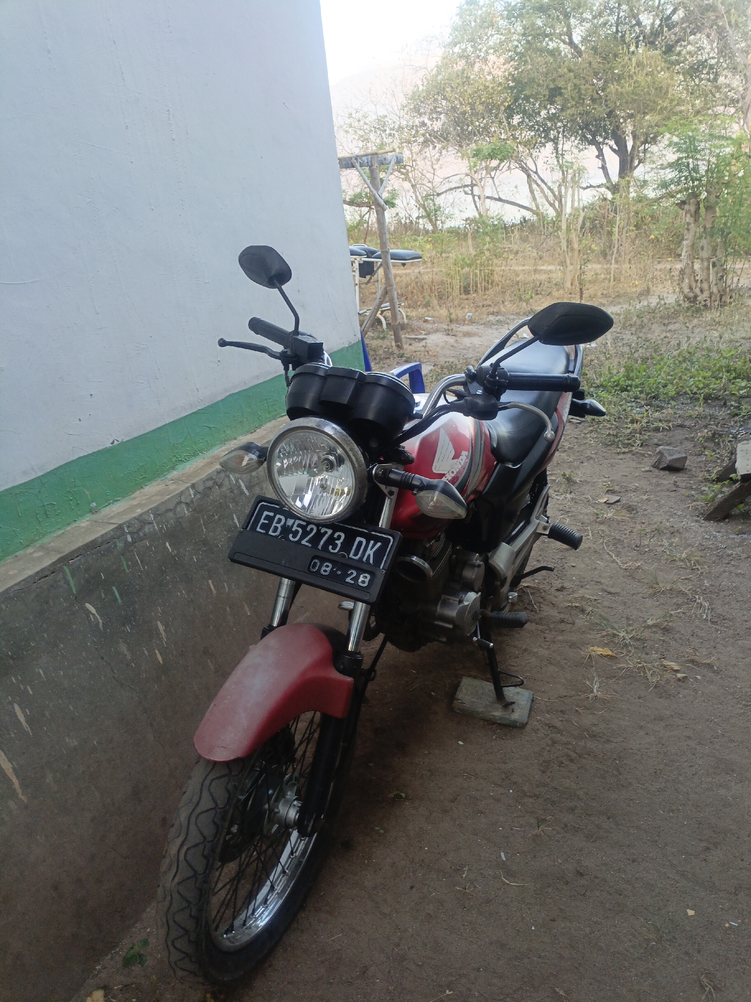 A red and black Honda motorcycle with license plate EB 5273 DK is parked on a dirt ground next to a white wall with a green stripe near the bottom. The motorcycle's front tire is partially visible, along with its headlight and handlebars. In the background, there are trees and dry vegetation. The scene appears to be outdoors in a rural or semi-rural area, possibly in Solor Barat, Indonesia, given the presence of the license plate. There are no people visible in the image.