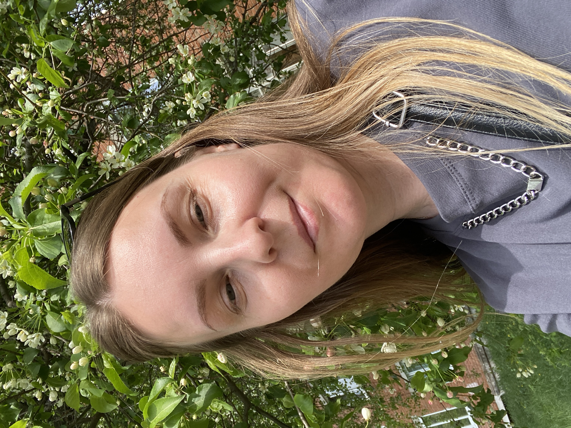 A woman with blonde hair, wearing a gray top with a silver chain detail, is posing in front of a tree with white blossoms and green leaves. The tree is located in a yard with a brick building visible in the background. It is difficult to determine the exact location in Noviy, Russia, from the image.