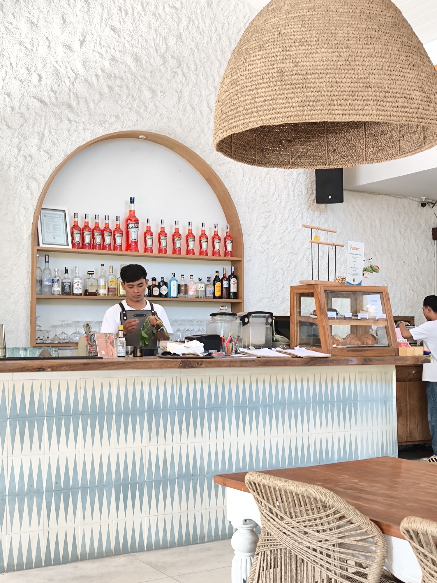 The image captures a scene inside a bar or restaurant, likely in Kuta, Indonesia. The bar is staffed by a young male bartender dressed in a white t-shirt and a dark apron, who appears to be preparing a drink, possibly garnishing it with greenery. Behind him, the bar shelves are stocked with numerous bottles, predominantly Aperol, with a variety of other spirits and alcoholic beverages.
The bar counter itself is adorned with a pattern of light blue and white geometric tiles, topped with a wooden countertop. To the right of the bartender, a display case filled with pastries and baked goods stands. Another individual, partially visible, is standing to the right near the display case.
In the foreground, a wooden table with rattan chairs suggests a seating area for customers. Above, a large, woven lampshade hangs from the ceiling, creating a warm and inviting ambiance. The walls are white with a textured finish, adding to the overall aesthetic of the establishment.