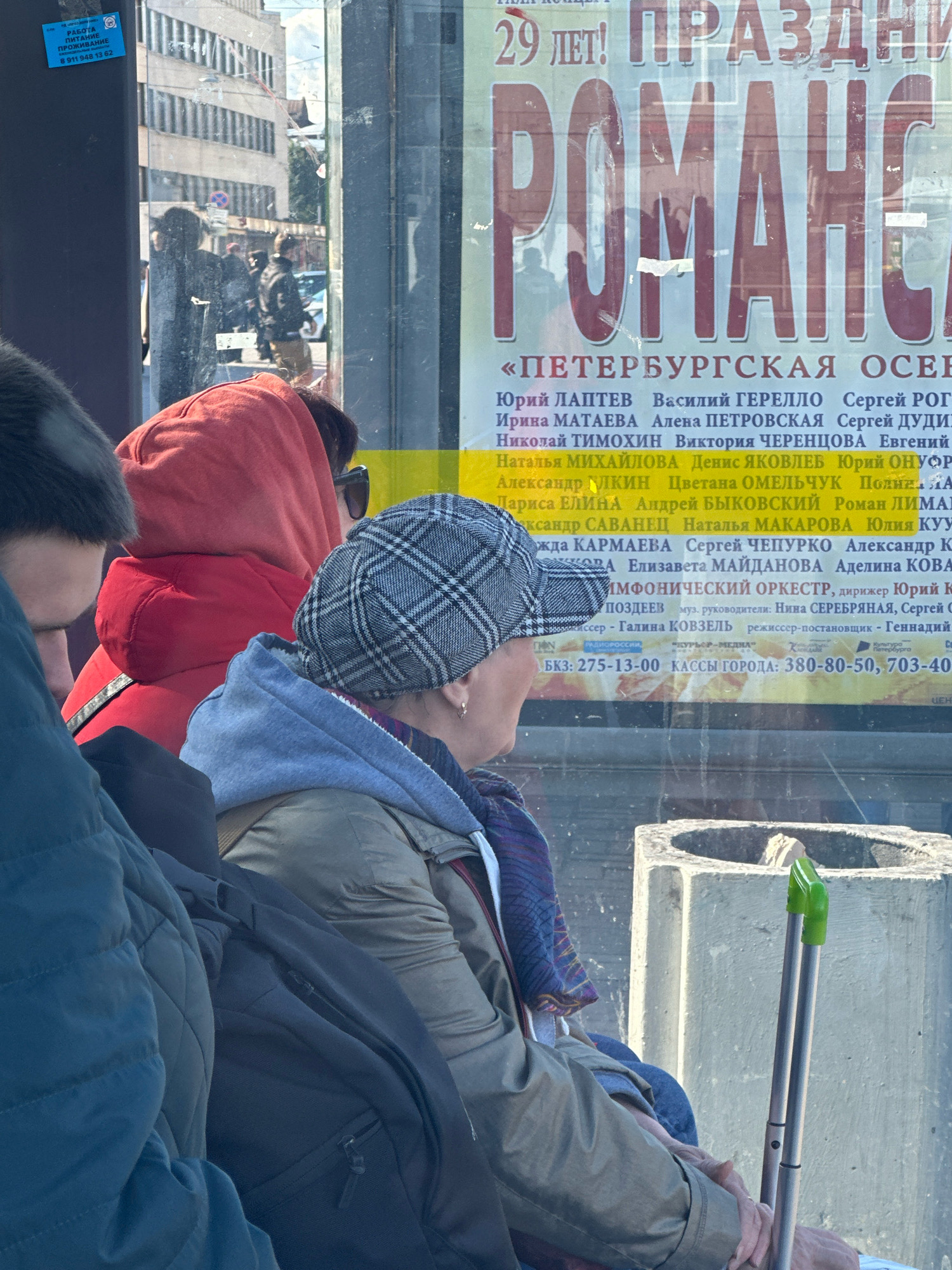 The image shows a group of people inside what appears to be a bus or tram, looking out a window at a street scene in Saint Petersburg, Russia. The window has advertisements on it, with large text in Russian. The people are wearing casual clothing, including a red hoodie, a plaid cap, and various jackets. Some are looking intently at the advertisement, while others are obscured from view. Outside the window, buildings and streetlights are visible, along with reflections on the glass. The overall lighting suggests it is daytime. A concrete structure, possibly a bench or bollard, is visible in the foreground, along with part of a foldable cane with green handles.