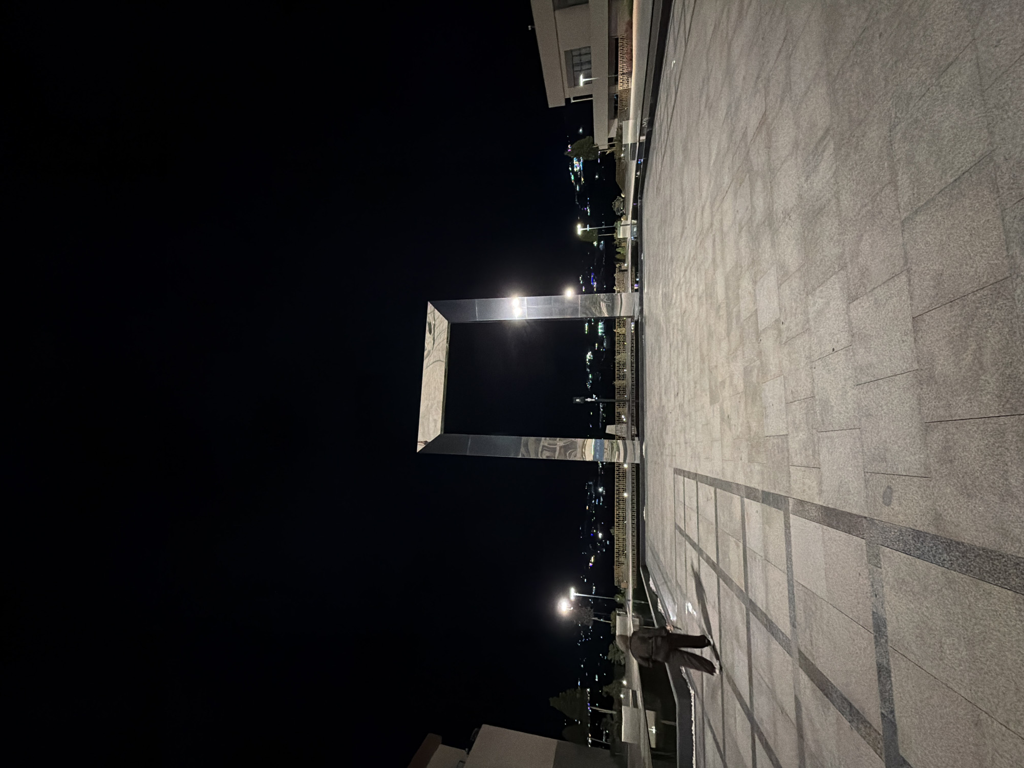 A person walks alone across a vast, paved plaza at night, heading away from the viewer and towards a monumental, arch-like structure. The plaza is illuminated by streetlights and the ambient glow of a city visible in the background, nestled amongst hills. The archway, made of a highly reflective material, stands prominently in the center of the plaza, its surfaces mirroring the night sky and the surrounding lights. To the left and right of the arch, modern buildings with clean lines and large windows are visible. The ground is paved with large, rectangular tiles arranged in a grid pattern. The overall scene suggests an open, public space in a city, likely designed for gatherings or as a landmark. The location is identified as Chinarkent, Uzbekistan.