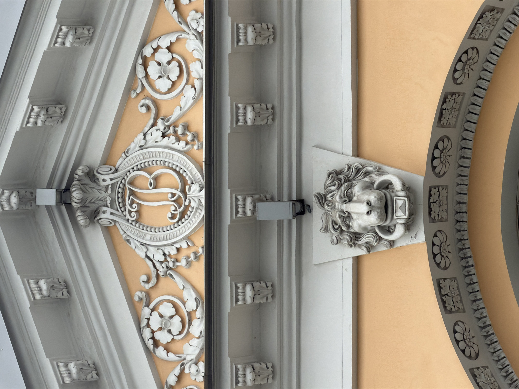 The image shows an ornate building facade in Unknown, Unknown. A beige-colored wall serves as the backdrop for various architectural embellishments. On the left, a gray, geometrically patterned structure with floral motifs is visible, along with a large decorative element featuring a script letter within a circular frame. In the center, a vertical section features stepped rectangular projections. To the right, a lion head sculpture is mounted on the wall, juxtaposed with a segment of a large, circular gray arch. The photograph is taken from a low angle, looking upwards.