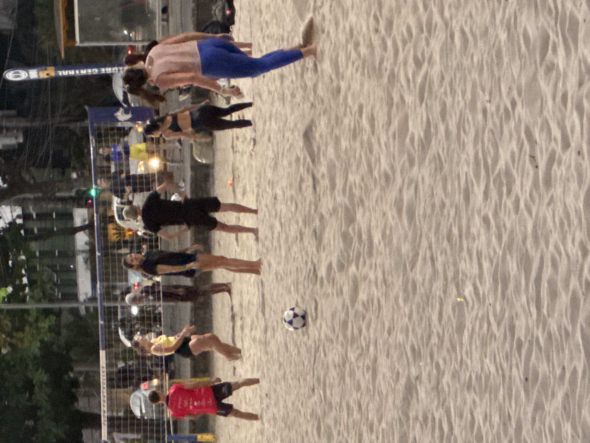 A group of people are playing soccer on a sandy beach, likely in Niterói, Brazil, given the context of the prompt. The scene appears to be during the day, as indicated by the lighting. There are at least seven individuals visible, engaged in the sport. One person is wearing a red shirt and black shorts, another in a yellow top and black shorts, and others in black tops and shorts or athletic wear. A soccer ball is prominently featured in the foreground. In the background, a volleyball net is set up, suggesting multiple sports activities are occurring. There are trees and buildings visible in the distance, characteristic of an urban coastal environment. The sand is rippled, indicating activity or wind. The overall impression is of a casual, recreational gathering.