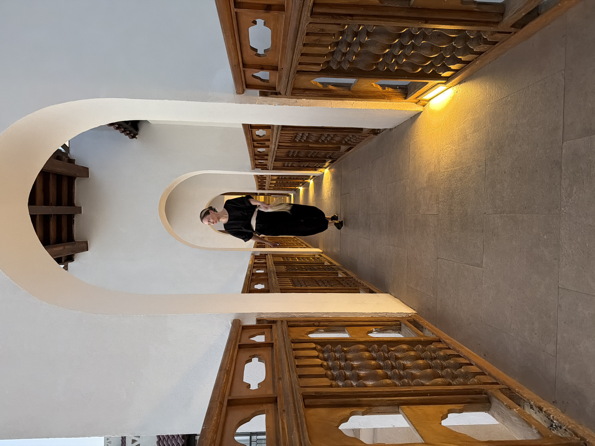 A photograph captures a woman standing in a gracefully curved hallway, characteristic of traditional architecture. The woman, positioned centrally, is dressed in a long-sleeved black garment, possibly an abaya or dress, and wears dark shoes. Her light-colored hair is pulled back, and her face is seen in profile as she looks forward into the corridor with a neutral expression.
The hallway is defined by multiple receding white arches, creating a tunnel-like effect and a strong sense of depth. Along both sides of the corridor, below the arches, are elaborate wooden balustrades or decorative panels. These panels are crafted from medium-brown wood, featuring intricate carvings, including wave-like motifs and distinctive keyhole-shaped cut-outs, which contribute to a culturally rich aesthetic.
The floor is made of light-colored, possibly stone or ceramic, tiles. Warm, yellow artificial lights are discreetly placed along the base of the walls on the right, casting an inviting glow upwards that highlights the textures of the carved wood and the tiled surfaces. The lighting suggests an indoor setting, possibly an evening atmosphere or a space designed for artificial illumination. The architectural style, with its arches and decorative woodwork, aligns with the context of Sharm El Sheikh, Egypt. No visible text is present in the image.
