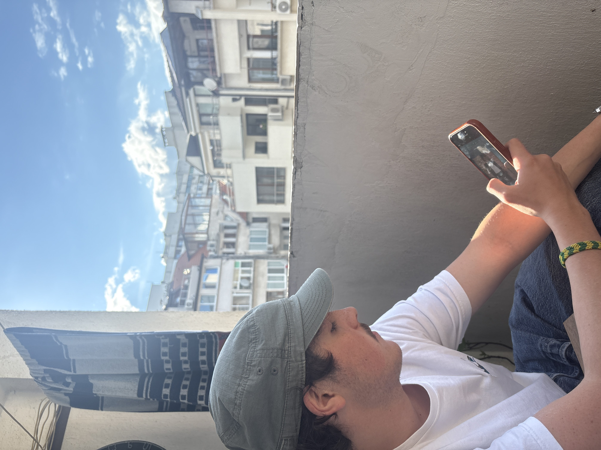 A young Caucasian male, wearing a white t-shirt, blue jeans, and a green cap, is seated on a balcony in Burgas, Bulgaria. He is looking down at his mobile phone, which he holds in his right hand. A woven bracelet is visible on his left wrist. In the background, several buildings with white facades and balconies are visible under a bright blue sky with scattered clouds. A striped curtain hangs on the left side of the frame. The sun is casting a warm light on the scene.