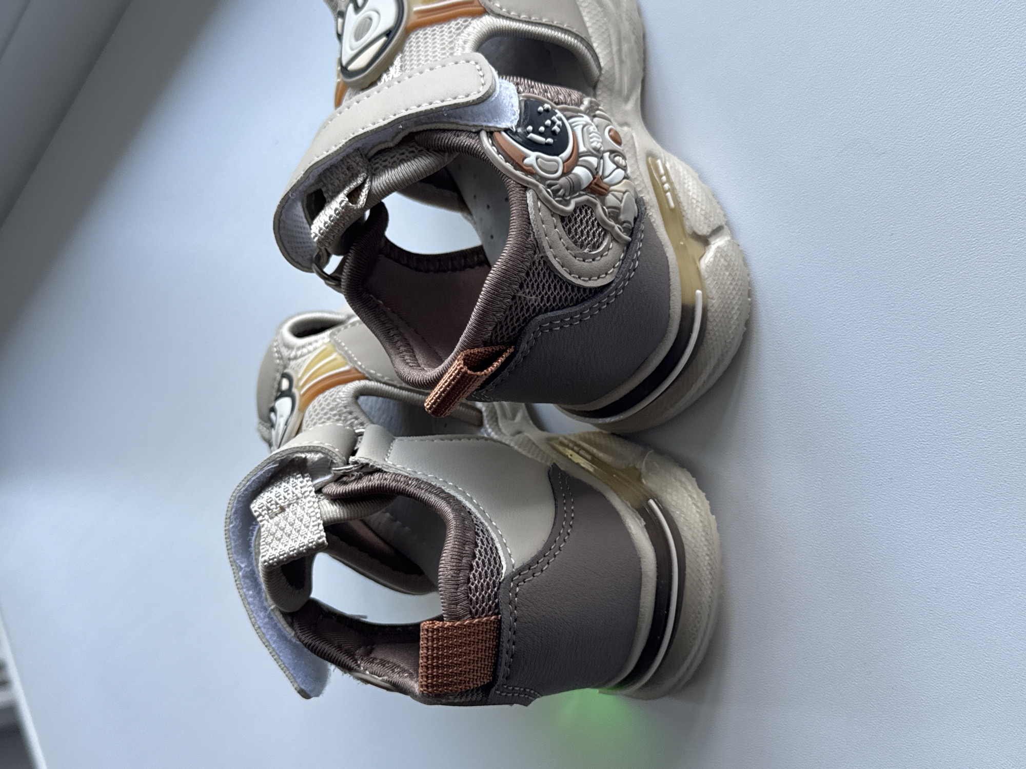The image displays a pair of children's sandals placed on a white, textured surface. The sandals are primarily beige and brown, with a design featuring an astronaut character. One sandal is positioned behind the other, both angled to show their sides and soles. The background is a pale blue, out of focus, suggesting an indoor environment. The lighting is bright, casting a slight green hue on the left side of the rear sandal. The scene is static, with no people or actions depicted. The specific location of Яромінскі сельскі Савет, Belarus, is not visually indicated in the image.
