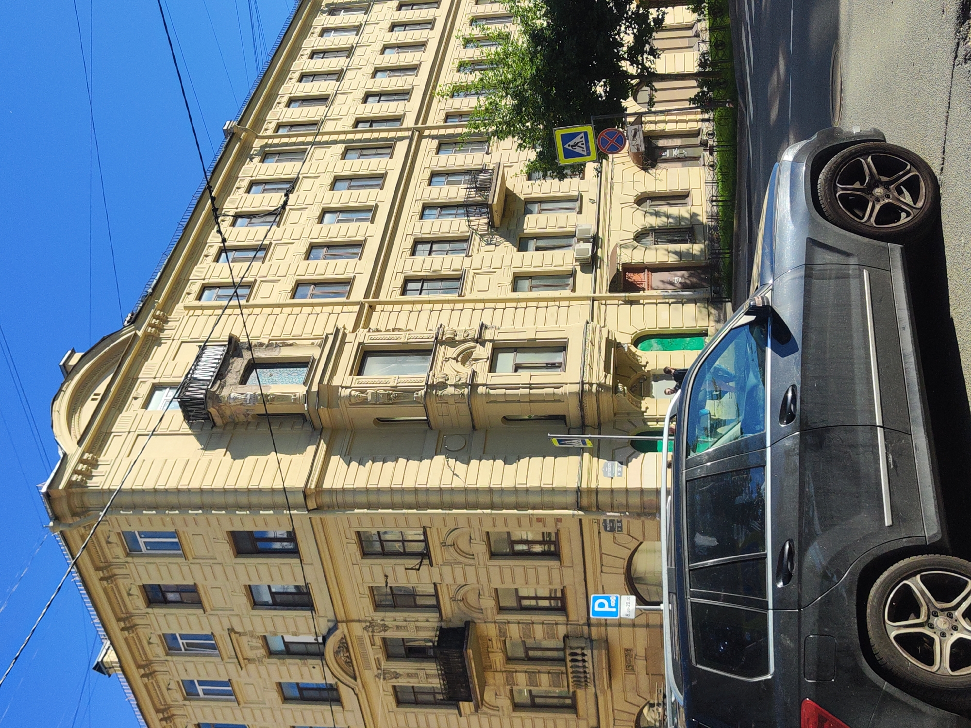 A dark-colored car is parked on a street next to a tall, yellow apartment building with ornate architectural details. The building has many windows, some with balconies. The scene is illuminated by bright sunlight, suggesting it is daytime. Overhead power lines are visible against a clear blue sky. Traffic signs are posted on the building, including a yellow diamond sign with a black border and a blue circular sign with a red border and a diagonal red bar. A blue parking sign with a white "P" is also visible. The car's wheel and part of its body are in the foreground. The overall impression is of an urban environment on a sunny day. The image is oriented vertically.