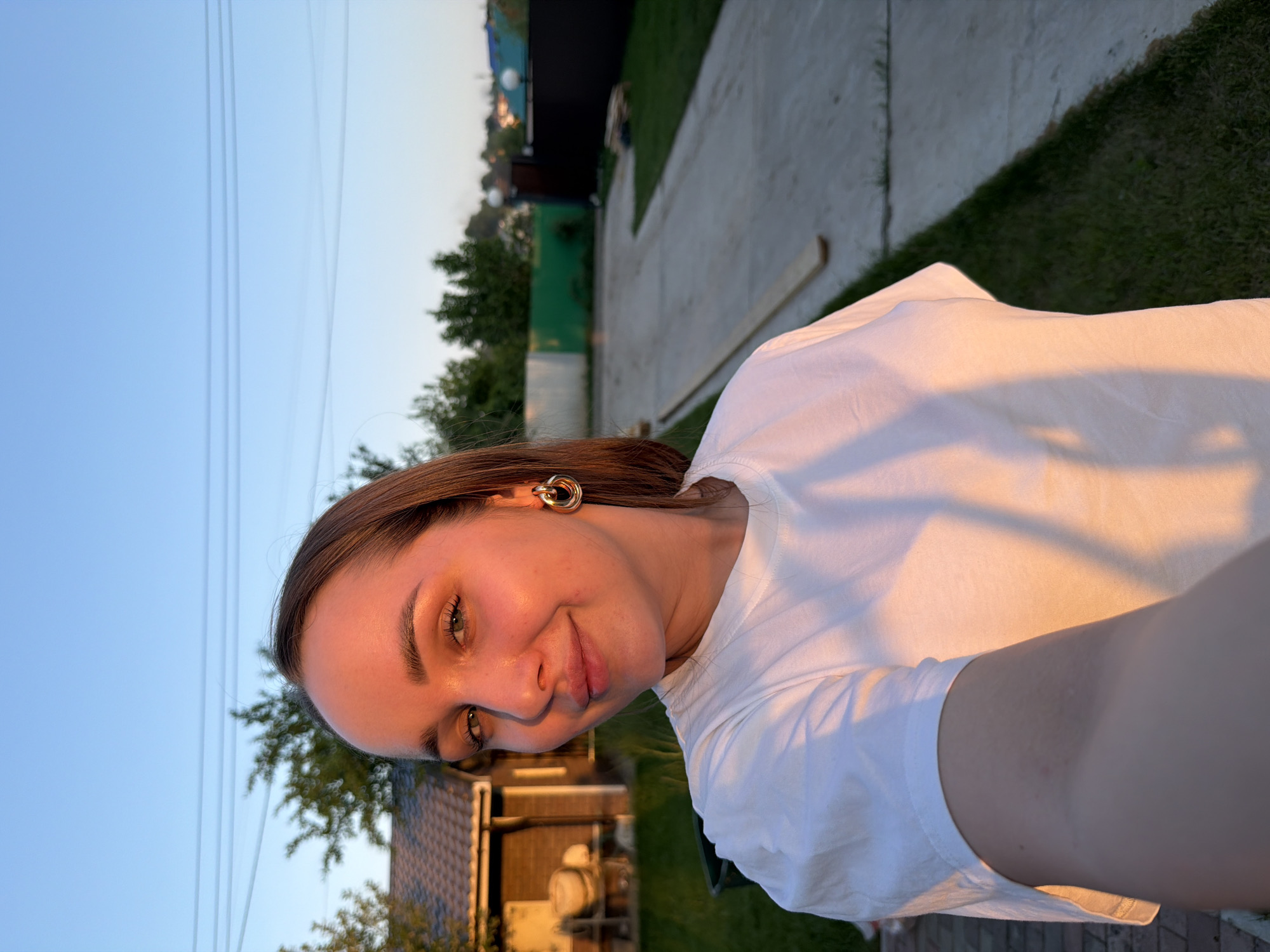A young woman is posing for a selfie outdoors, bathed in warm, golden light, likely during sunset or sunrise. She has fair skin, light brown hair pulled back, and is wearing a white t-shirt and distinctive gold earrings. The background includes a blue sky with power lines visible, trees, a wooden building with a tiled roof, a concrete walkway, and green grass. The location appears to be a residential area on the outskirts of Chaikovsky, Russia.