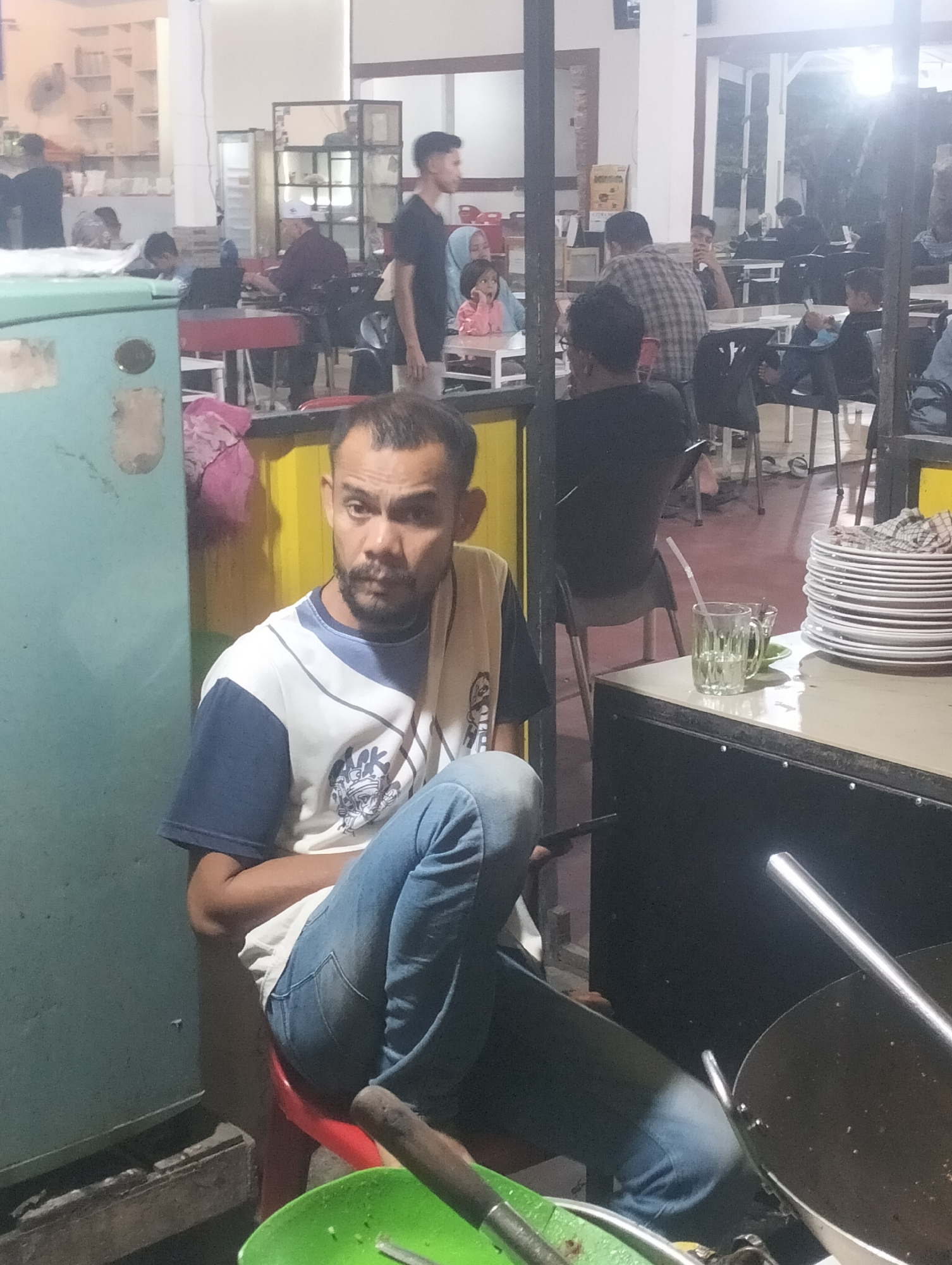 In the city of Lhokseumawe, Indonesia, a man is seated in the foreground. He is wearing a blue and white t-shirt and blue jeans. His facial features include a mustache and a goatee. He is sitting on a red stool. To his right, a stack of white plates is visible on a black surface, next to a glass of water with a straw. Behind him, a turquoise refrigerator stands to the left. Further in the background, patrons are seated at tables in what appears to be an eatery or cafe. Some individuals are visible in the middle ground, including a person in a checkered shirt and a person with a white cap. The environment is dimly lit, suggesting it may be evening or indoors with artificial lighting.