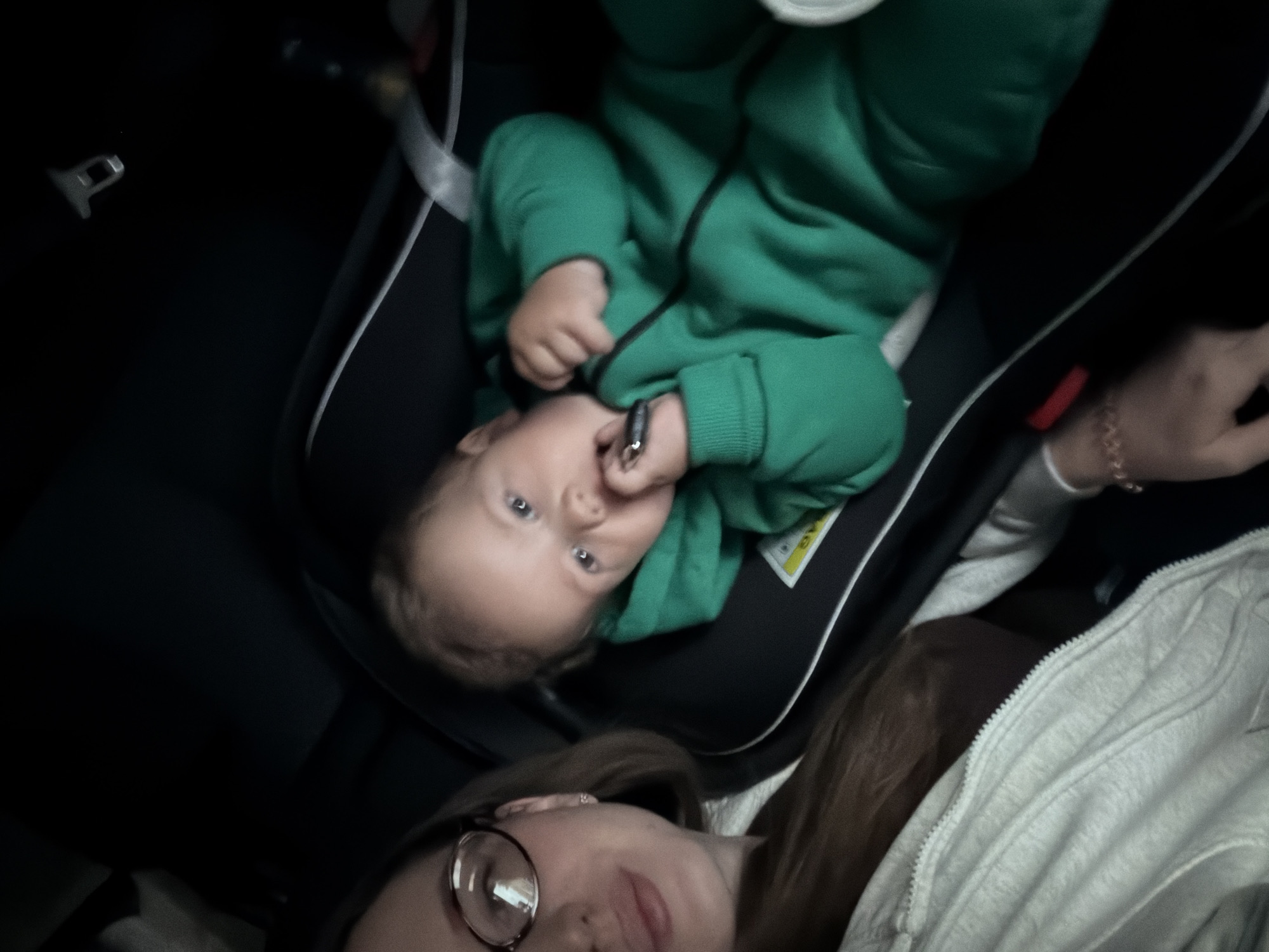 A close-up, high-angle selfie shows a woman and a baby in a car. The woman, wearing glasses and a light-colored jacket, is in the upper portion of the frame, looking down. The baby, dressed in a green jumpsuit, is in a car seat in the lower part of the frame, facing the camera and putting something in its mouth. The background is dark, suggesting the interior of a car. The scene appears to be taking place in Kazan, Russia, although this cannot be confirmed from the image alone.