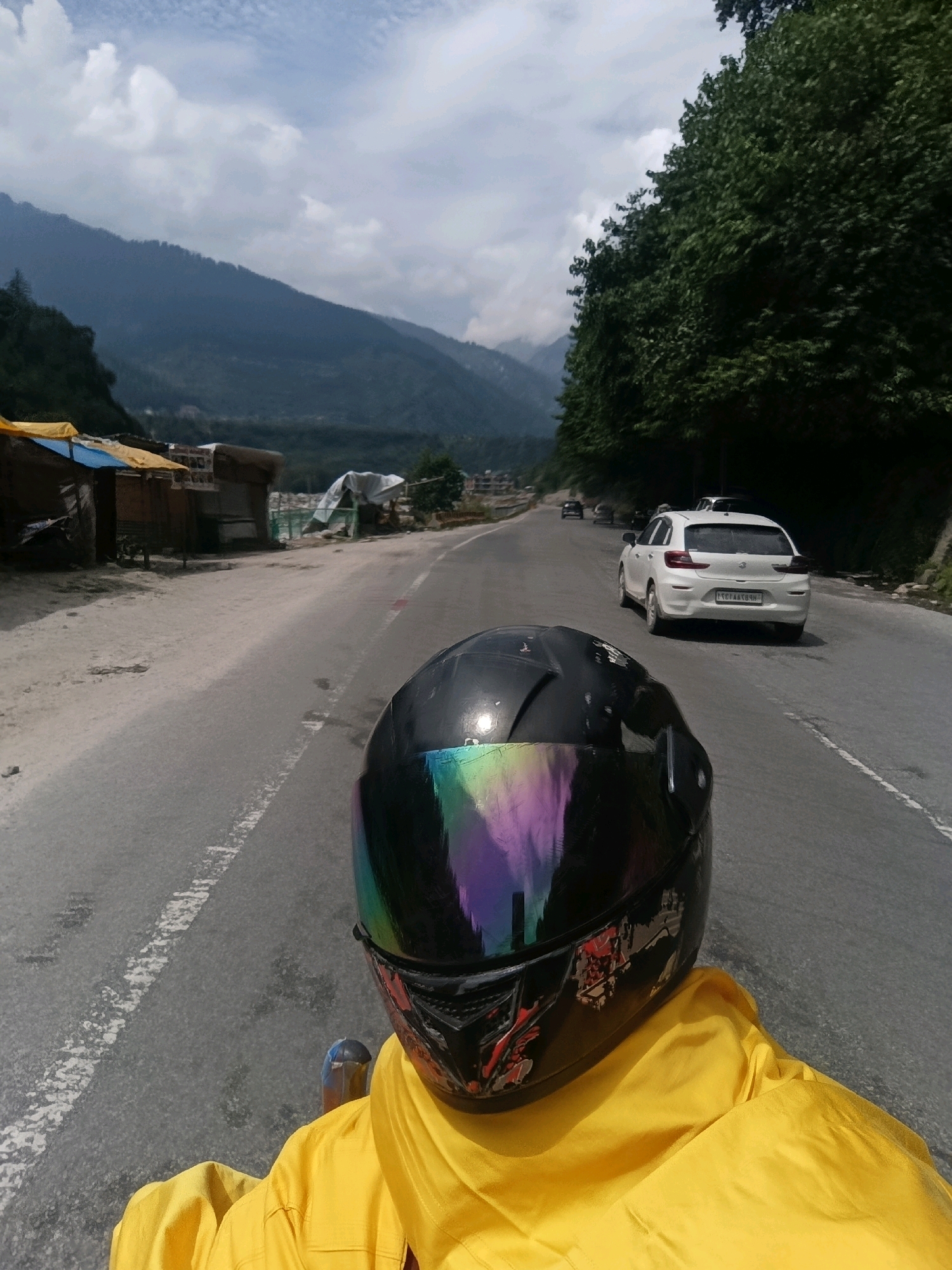 A first-person perspective shot shows a motorcyclist in a yellow jacket and a black helmet with a rainbow-colored visor on a paved road. The road is lined with trees on the right and some structures on the left. In the background, mountains rise, shrouded in mist. A white car is visible on the road ahead, along with other vehicles and some tents. The scene appears to be in Kalath, India.