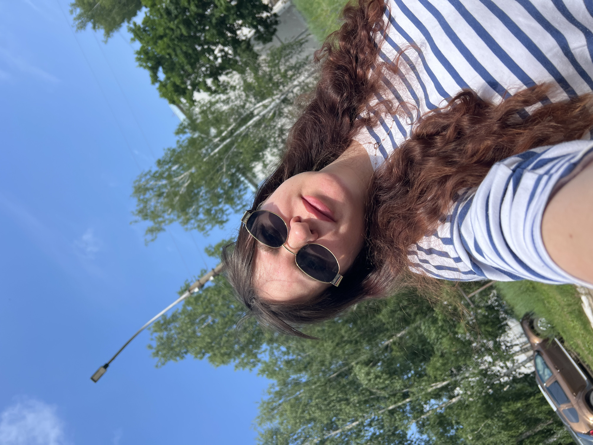 A woman with long, wavy, reddish-brown hair and sunglasses is taking a selfie outdoors on a sunny day. She is wearing a blue and white striped t-shirt and a delicate rose gold necklace. The background shows a bright blue sky with a few wispy clouds. To the left, a tall lamppost is visible. Further back, green trees and a white building can be seen. In the lower left corner, a brown SUV is parked on a grassy slope. The image appears to have been taken in a residential or park area. The location is Noviy, Russia.