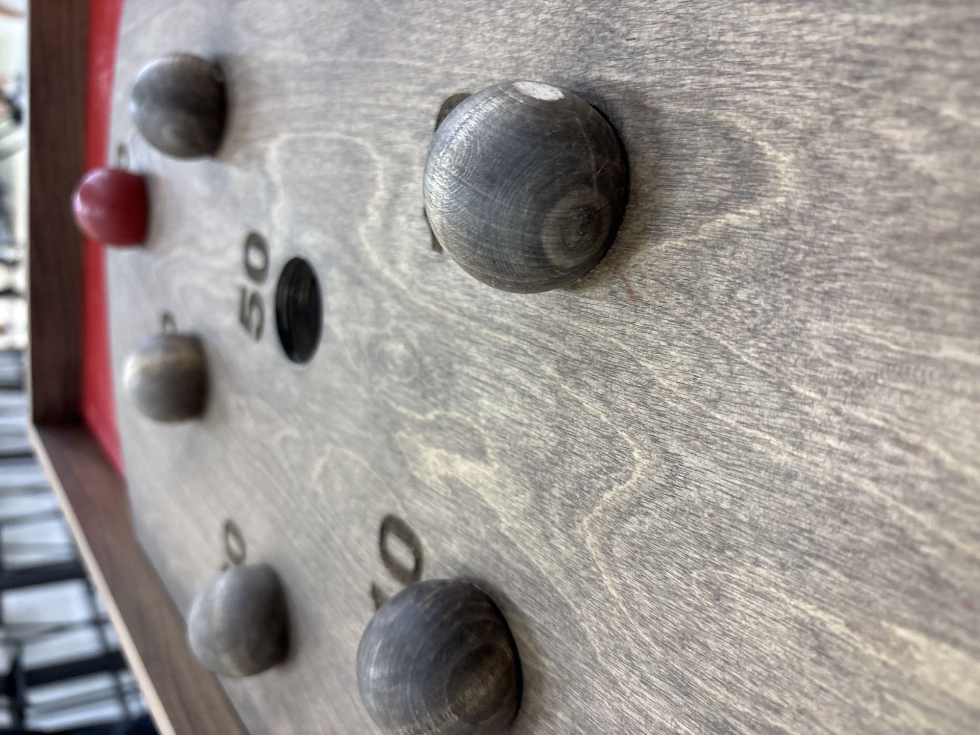 A close-up, low-angle shot shows wooden balls on a wooden surface with numbers and holes. In the foreground, a dark wooden ball is positioned on the right side, with some white paint smudged on its surface. To the left and slightly behind it, another dark wooden ball is visible. Further back, another dark wooden ball is seen in the upper left quadrant. In the middle ground, a dark wooden ball is positioned near a hole labeled "50". Behind this, there are three more wooden balls, one greyish and two that are more rounded, with one red ball in the upper right. The surface appears to be part of a game board, with red fabric visible beneath the wooden surface in the top right corner. The scene is lit from above, creating shadows. There are no visible people or signs of the city of Orenburg, Russia.