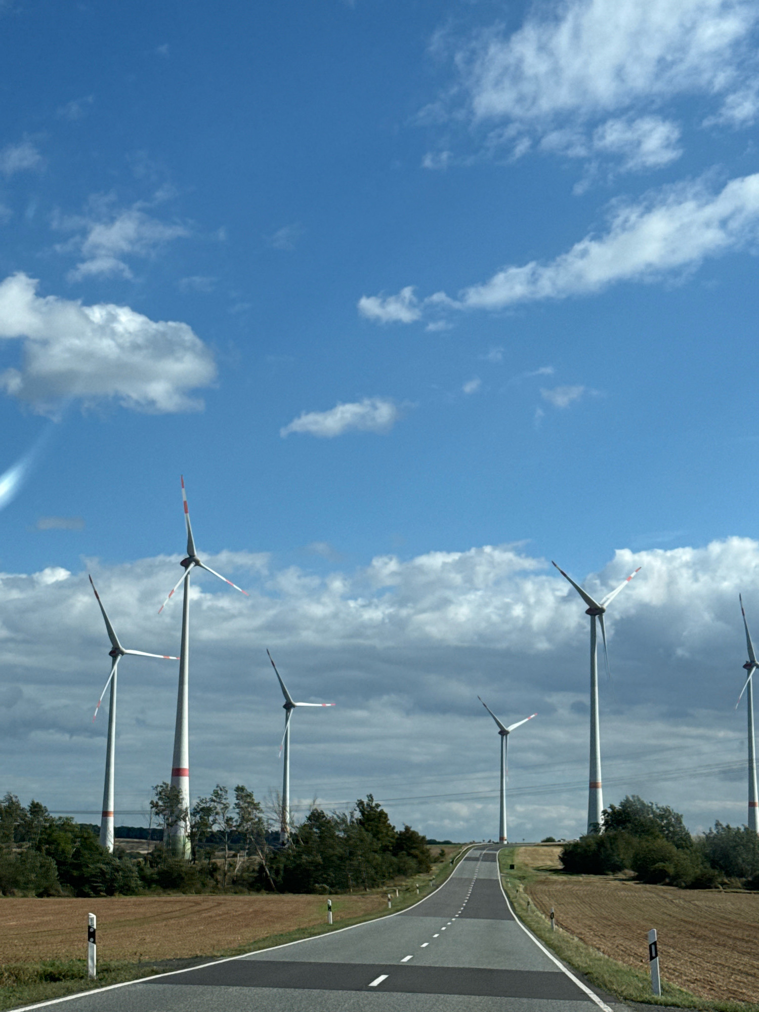 The image depicts a view of wind turbines in the distance, with a paved road leading towards them. The sky is blue with scattered white clouds. The foreground features the road with white dashed lines marking the lanes and reflective markers on the side. To the left and right of the road are fields of dry grass or crops, and some trees and vegetation. Several tall wind turbines with white blades and red tips are visible on a slight incline behind the fields. The scene appears to be in a rural area, possibly in or around Kleinfurra, Germany, given the context. There are no people or animals visible in the image. The overall impression is one of a clear, sunny day in a landscape dominated by renewable energy infrastructure.