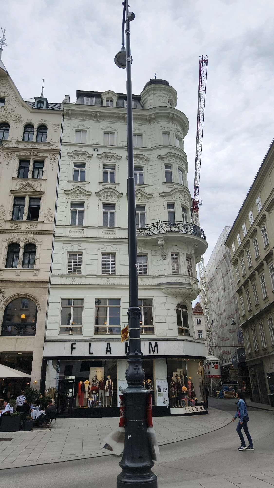 The image depicts a street scene in Vienna, Austria, under an overcast sky. In the foreground, a dark lamppost stands prominently. To the left of the lamppost, a building with ornate white facades and numerous windows is visible. Below the windows, a shop with the name "FLAMM" is displayed in large black letters. The shop window showcases mannequins dressed in clothing. An outdoor seating area with tables and chairs is set up on the sidewalk to the left of the shop, with a few people seated.
To the right of the lamppost, another building with a pale green facade and rounded corners dominates the view. This building also features multiple windows and decorative architectural elements, including a balcony. Further to the right, a tall construction crane is visible, indicating ongoing construction or renovation work. Adjacent to the pale green building, another building is partially obscured by scaffolding and white sheeting, also suggesting construction. The street itself appears to be paved with light-colored tiles, and a single person, wearing a denim jacket and dark pants, is walking across the street from right to left in the mid-ground. The overall impression is that of a busy urban environment with both historical architecture and modern development.