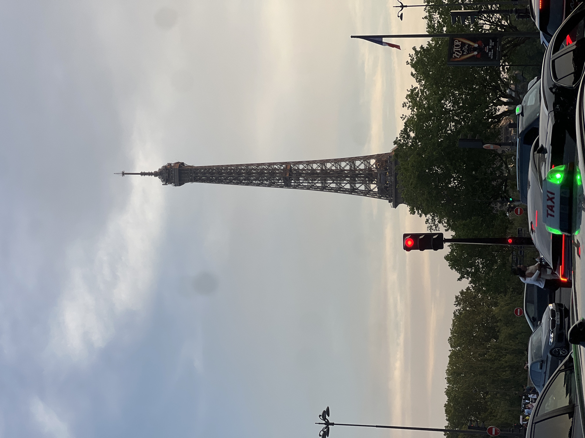 This image depicts a street scene in Paris, France, with the Eiffel Tower prominently featured in the background. The sky is overcast, with light and dark clouds present. The Eiffel Tower, a tall iron lattice structure, is visible towards the right side of the frame. In the foreground, there is heavy traffic, with several cars and taxis on the road. One taxi has a bright green light on its roof, indicating it is available. A traffic light is visible and is red. A couple is standing near the traffic light, with one person embracing the other. Trees line the street, and a French flag is planted on a pole near the Eiffel Tower. A poster for a ZZ Top concert is also visible on a pole. The overall scene suggests a busy urban environment during daylight hours, possibly late afternoon or early evening given the lighting.