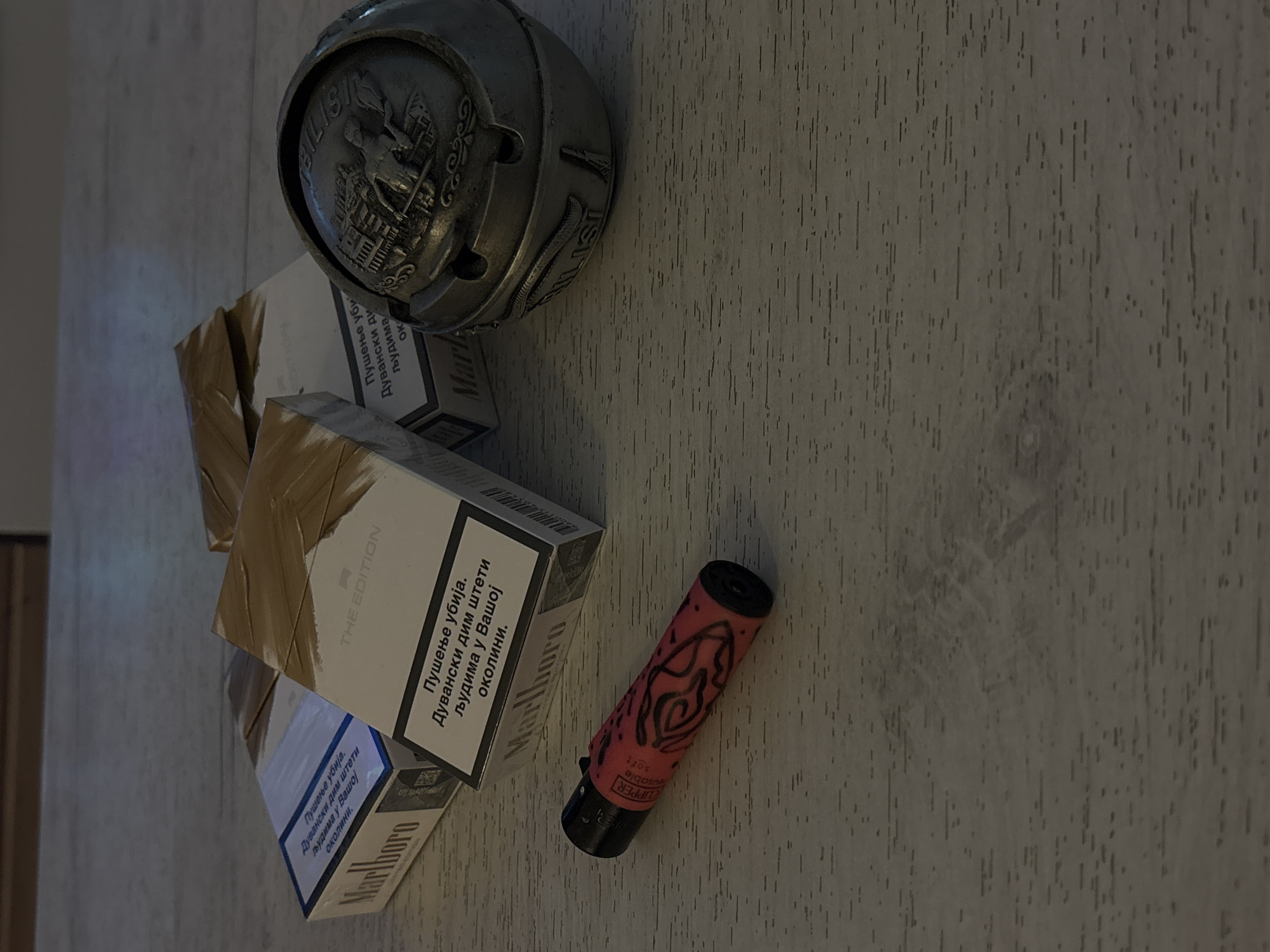 The image shows a collection of items on a textured, wooden surface, likely a table or countertop. The primary subjects are several packs of Marlboro cigarettes, some of which are partially wrapped in brown paper or tape, suggesting they might be new or stored. One pack prominently displays "THE EDITION" and features warning text in Cyrillic script that translates to "Smoking kills. Tobacco smoke harms you and people around you."
To the left of the cigarette packs is a distinctive, antique-looking metal object, possibly a decorative ashtray or a small container with an embossed crest featuring a building and a figure. The crest appears to have text around its circumference, with "TBILISI" clearly visible.
On the right side of the image, a red disposable lighter with a black top is placed. It has a pattern drawn on it, appearing to be a stylized floral or tribal design in black. The lighter is a "Clipper" brand, as indicated by visible text.
The overall scene is indoors, with soft lighting that casts subtle shadows, creating a somewhat moody atmosphere. The wooden surface has a dark, gray-brown color with a distinct grain pattern and appears to be a common household or commercial setting. The arrangement of items suggests they are casually placed, rather than part of a staged display. The presence of cigarettes and a lighter implies a context related to smoking. There are no people visible in the image.