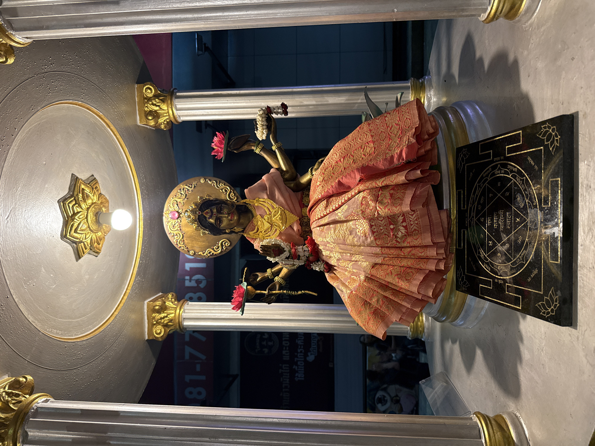 The image depicts a golden statue of a Hindu deity, possibly Lakshmi, adorned with jewelry and a pink and gold sari, positioned within a shrine-like structure. The structure features silver columns with ornate gold capitals, supporting a circular, grey niche with a gold trim and a bulb lamp at its center. A black plaque with gold Sanskrit inscriptions is placed beside the statue.
Outside the shrine, a glimpse of a Bangkok city street at night is visible. A portion of a business establishment is seen with Thai writing and numbers, suggesting the location is likely within the city. People are seen in the blurred background, indicating the shrine is in a public or semi-public space.